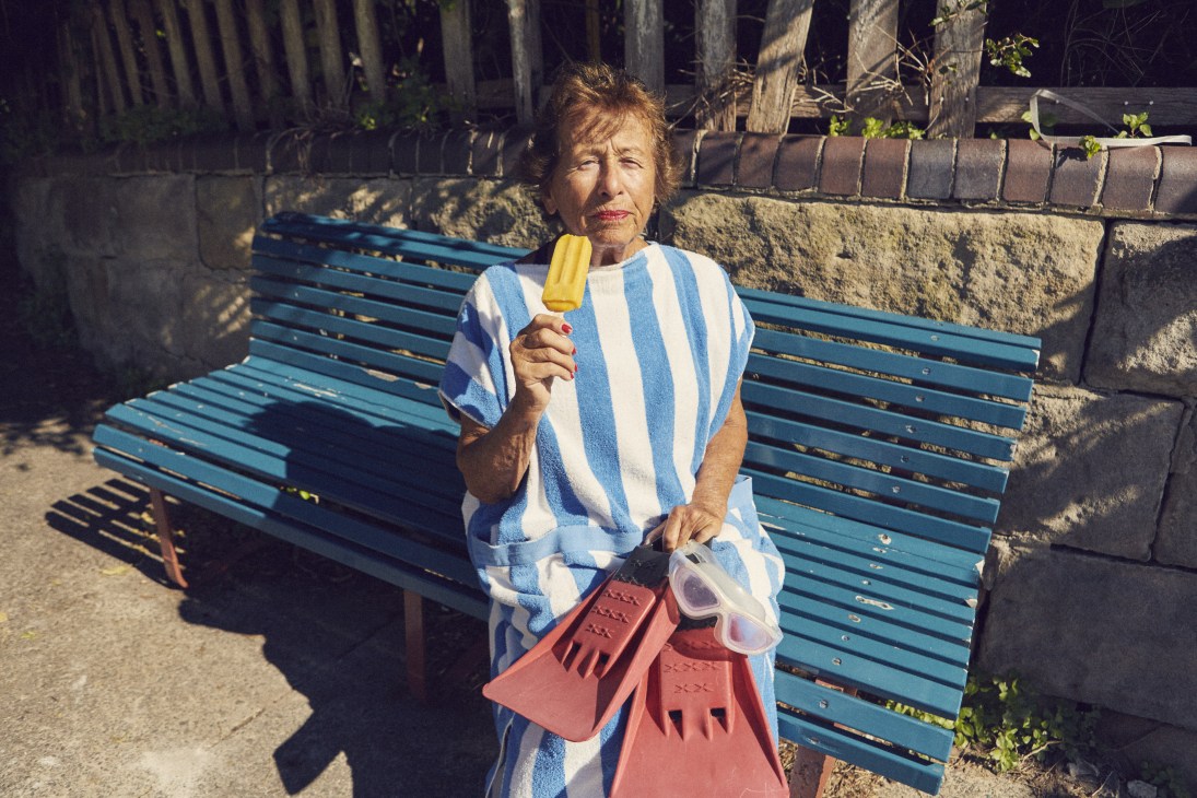 Woman in blue and white striped shirt sitting on a bench holding a yellow ice lolly and red beach bucket.