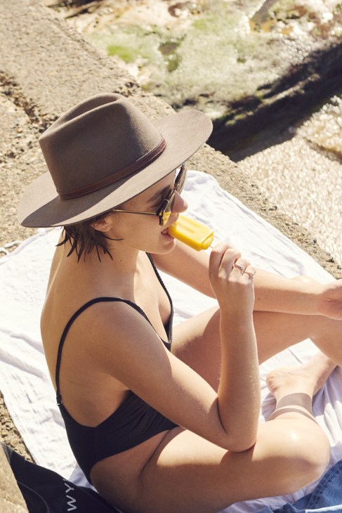 Woman in cowboy hat and sunglasses drinking yellow beverage while sitting on rocky ground.