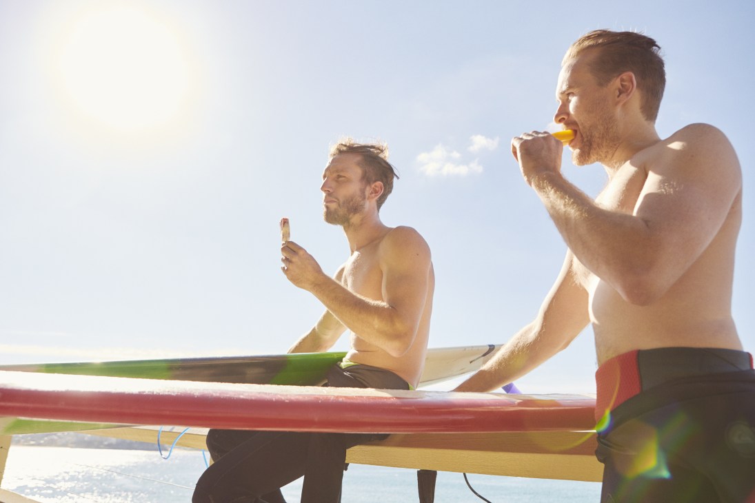 Two shirtless men eating ice lollies while sitting on surfboards in bright sunshine.