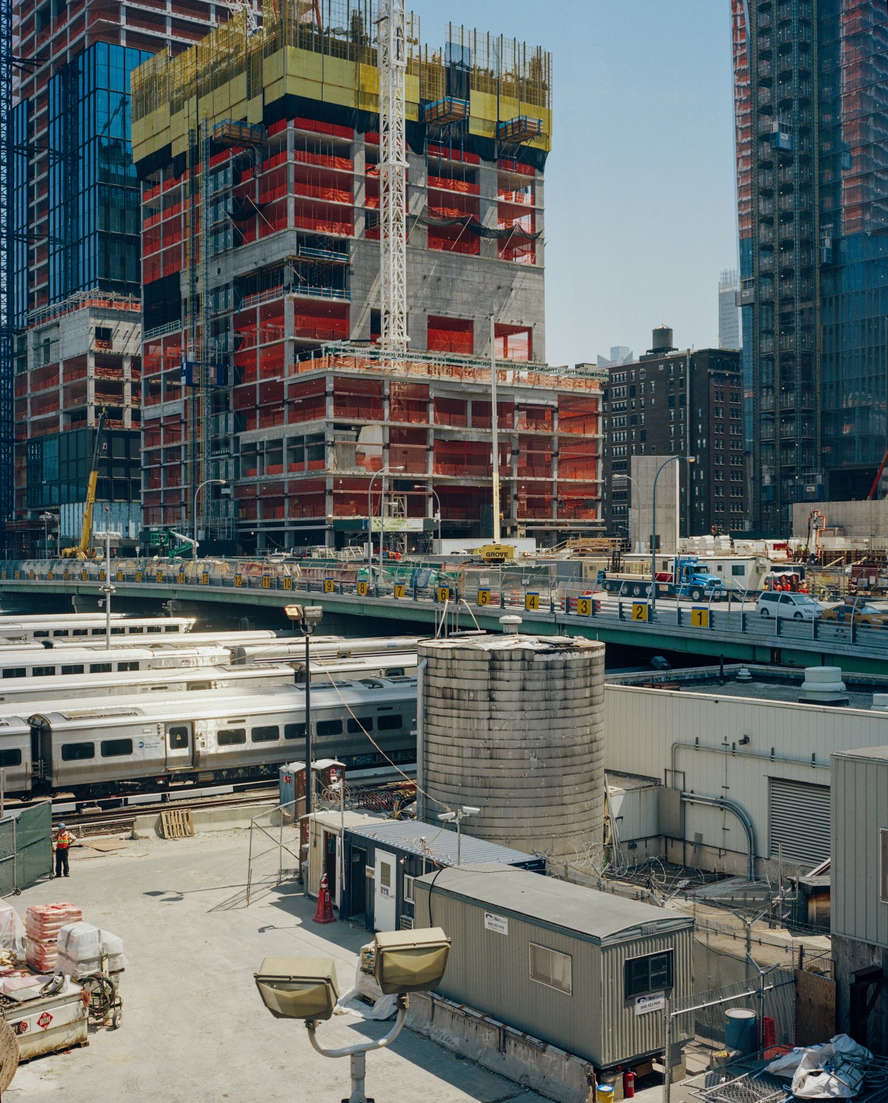 Construction site of Hudson Yards development with red-wrapped building, crane, and Manhattan skyline.