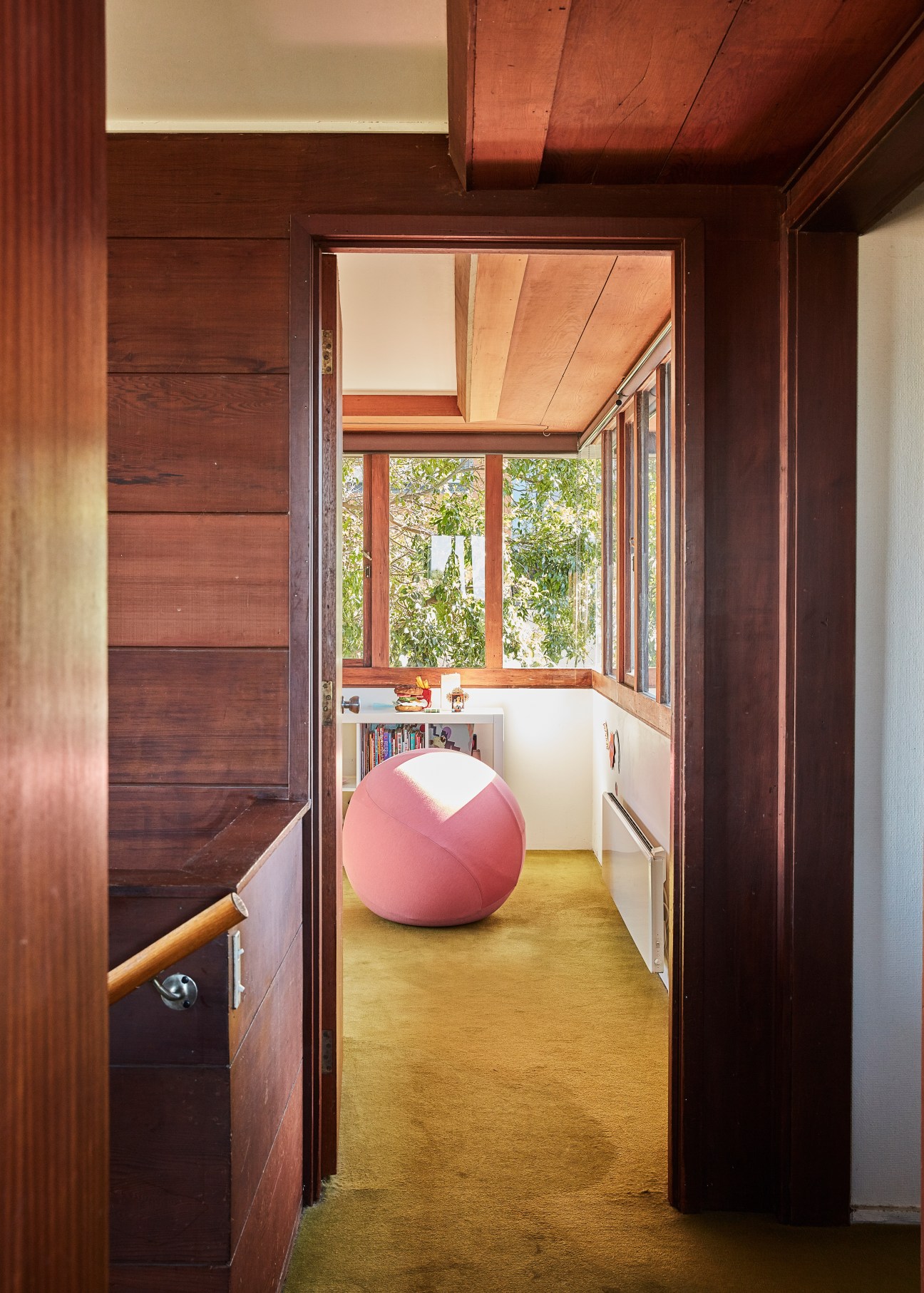 A modernist study with wood-paneled walls, a pink exercise ball, yellow flooring, and large windows framing trees outside.