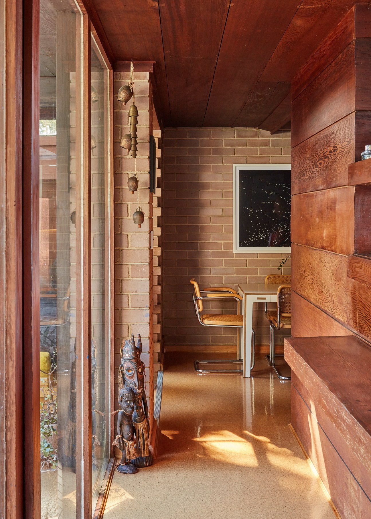 Narrow brick corridor lined with wood paneling and hanging bronze bells, featuring modernist furniture and artwork.