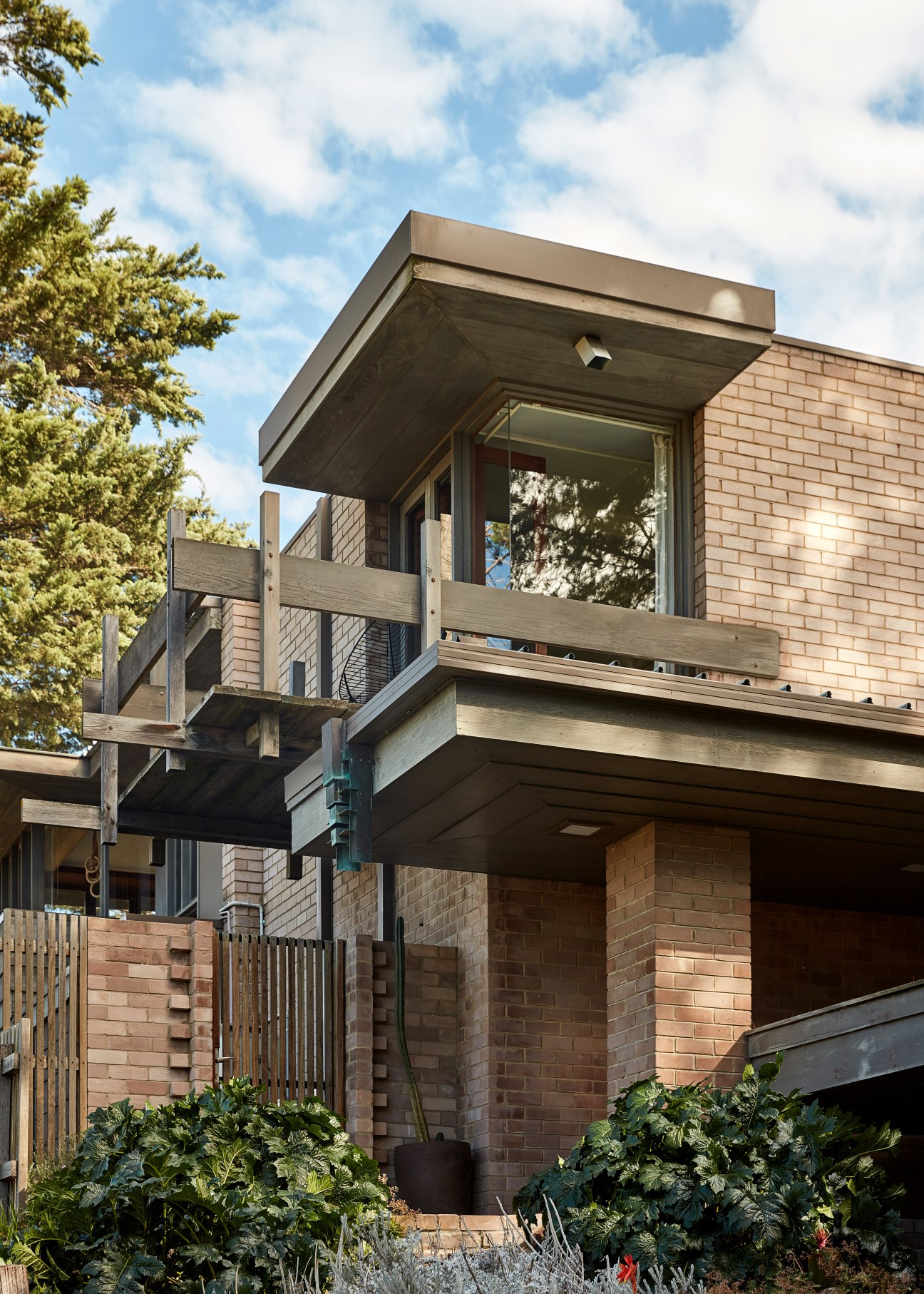 Modern brick residential building with cantilevered upper level and wooden railings, surrounded by green vegetation.