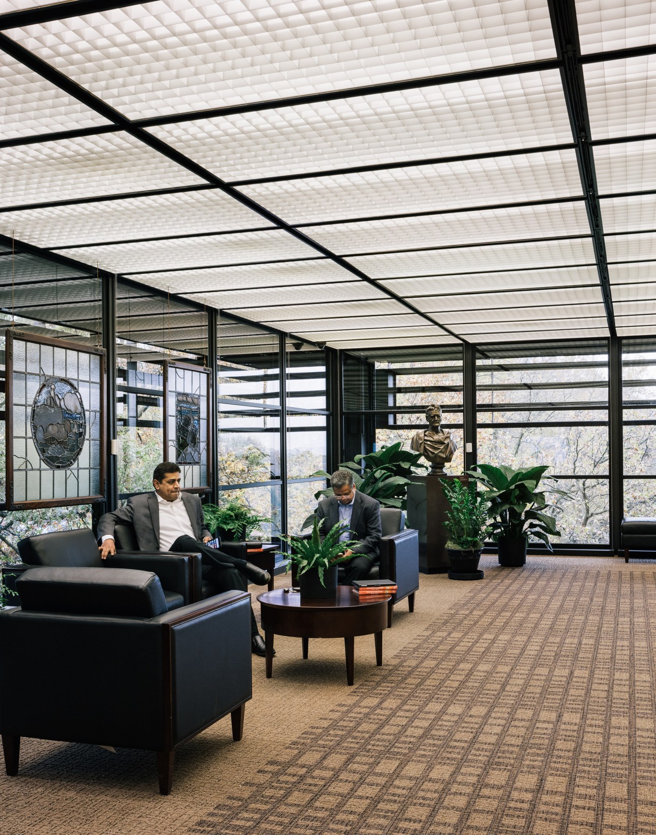 Two men in business attire sit in a modern office space with floor-to-ceiling windows, potted plants, and a geometric ceiling with black metal framing.