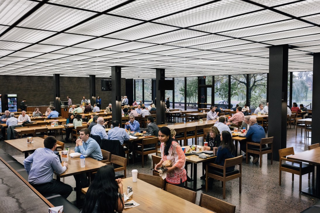 John Deere headquarters cafeteria with employees dining at wooden tables under a geometric ceiling and large windows overlooking the landscaped grounds.