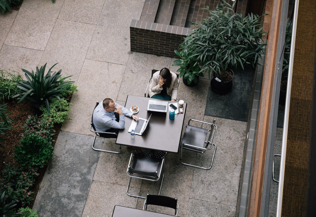 Two employees working at a table in a secluded outdoor meeting area surrounded by potted plants.