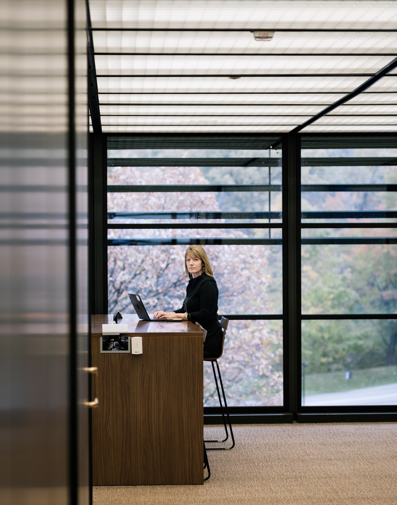 Woman working at desk in minimalist office space with floor-to-ceiling windows overlooking rural landscape.