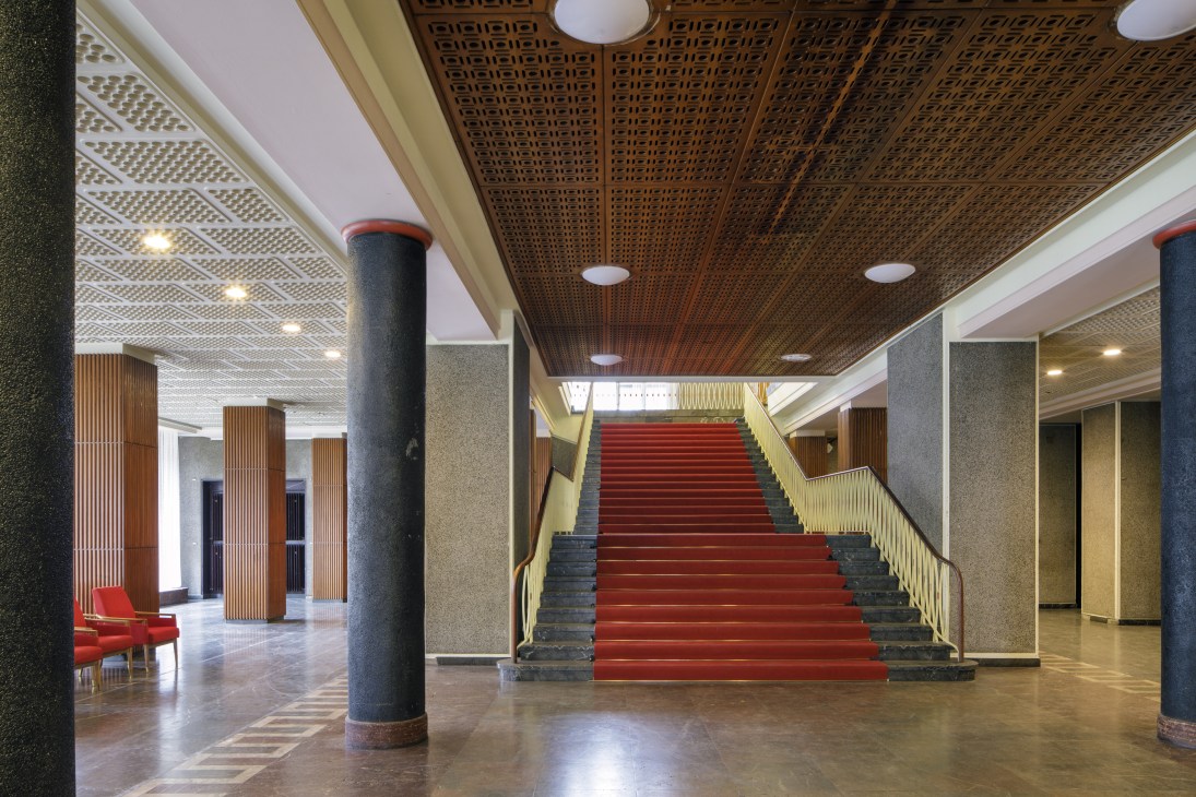 Red-carpeted staircase in the Funkhaus Block B foyer with geometric patterned wooden ceiling and supporting columns.