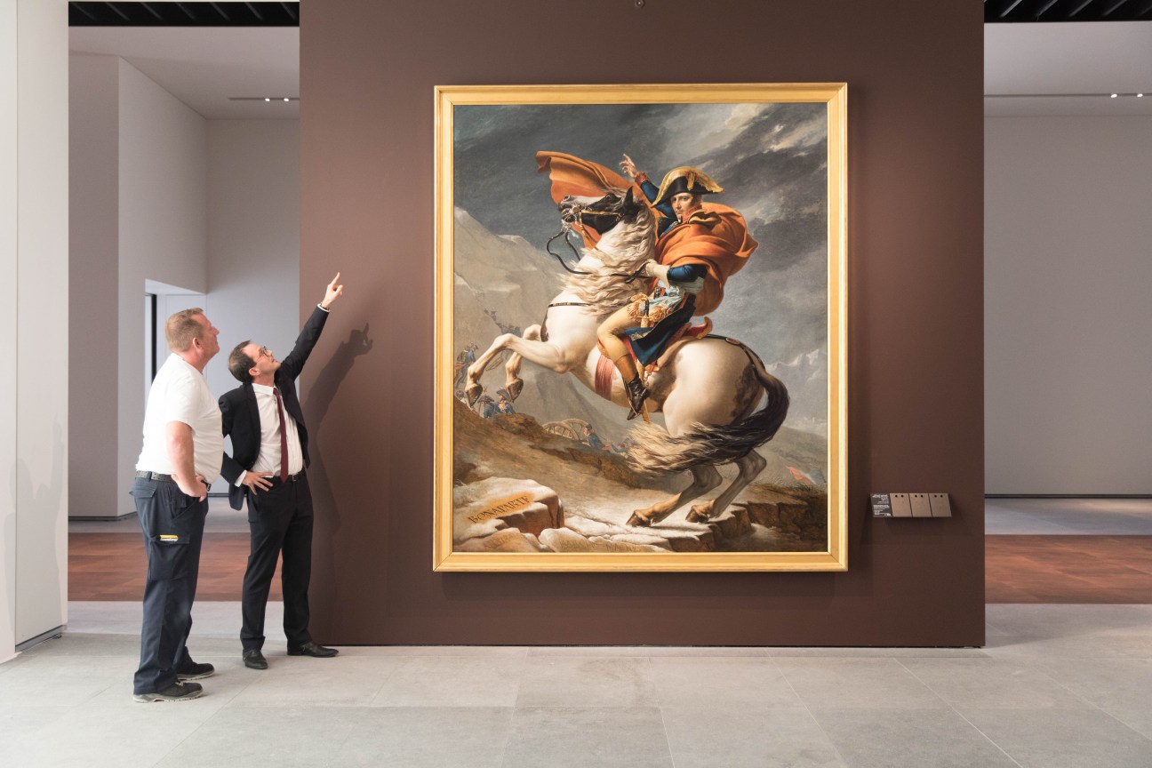 Two men pointing at Jacques-Louis David's 'Napoleon Crossing the Alps' hanging on a brown gallery wall.