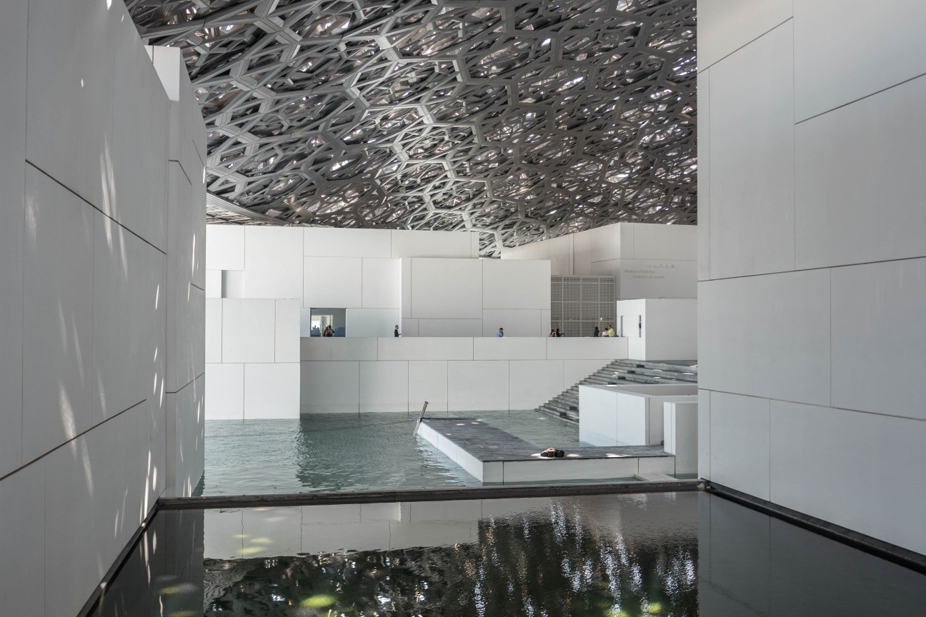 Interior atrium of Louvre Abu Dhabi with latticed roof, white walls, and water feature reflecting the geometric ceiling design.