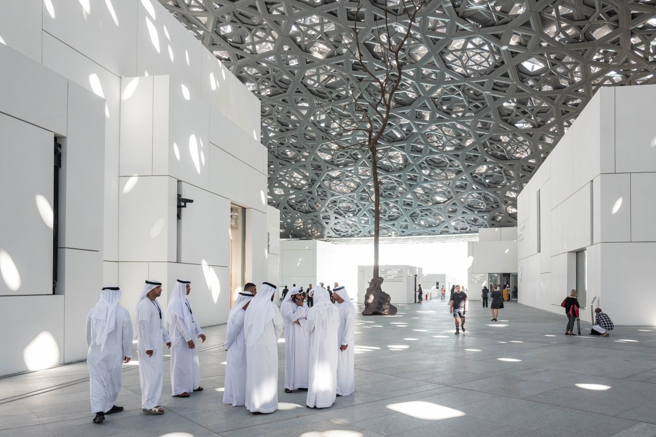 Louvre Abu Dhabi's atrium with Jean Nouvel's geometric lattice-work dome casting patterned shadows on white walls and floor, with visitors in white traditional dress.