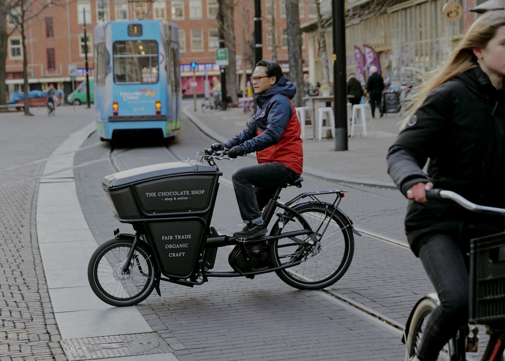 A cyclist riding a black cargo bike with "The Chocolate Shop" branding in an urban pedestrian plaza.