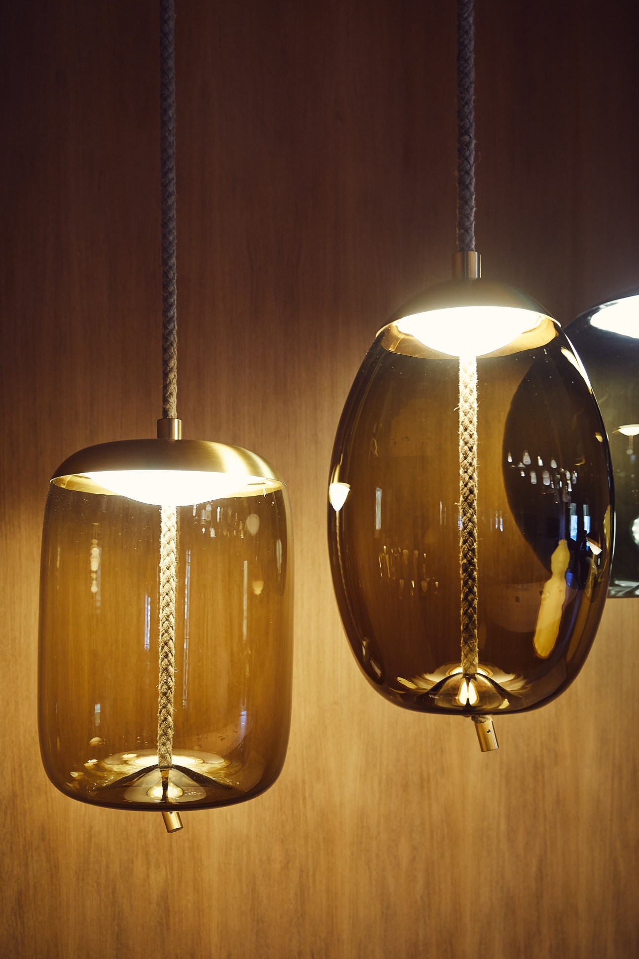 Two amber glass pendant lights by Brokis hang from textured cords against a warm wooden wall.