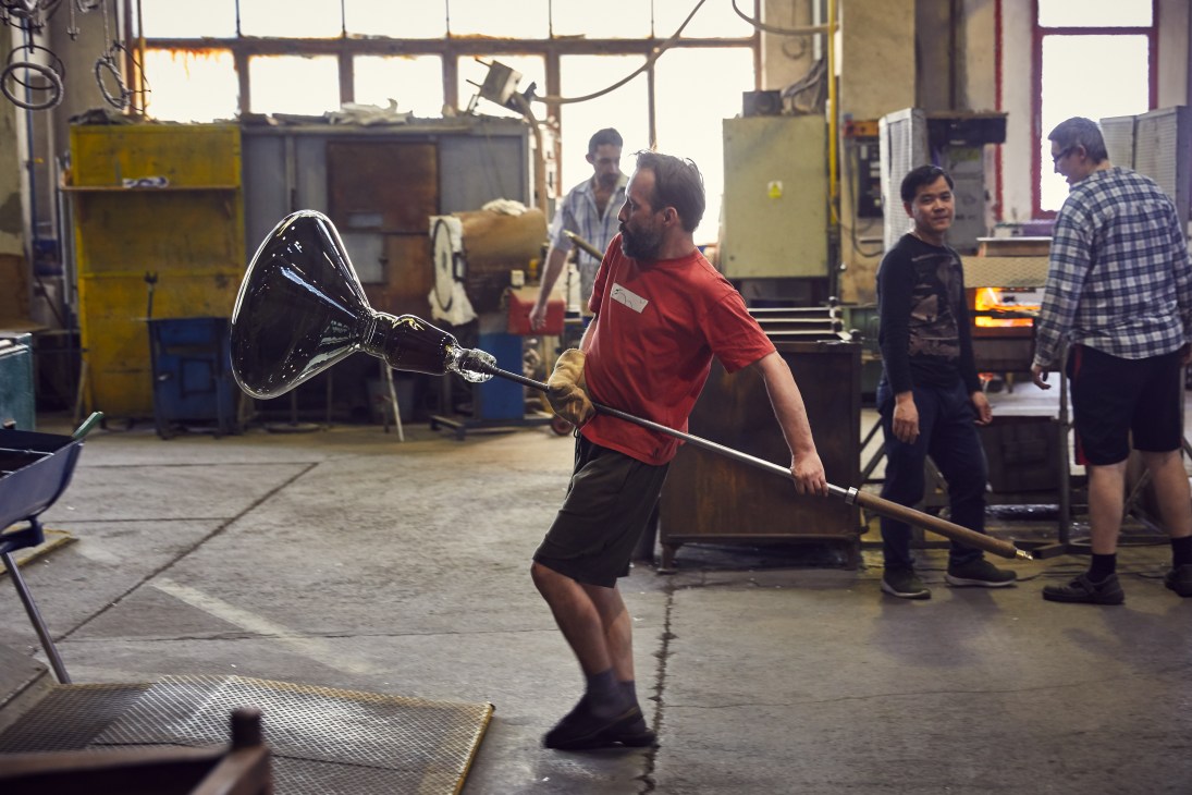 Glassblower in red shirt shapes molten glass using a blowpipe in a factory at Jenstejn Glassworks.