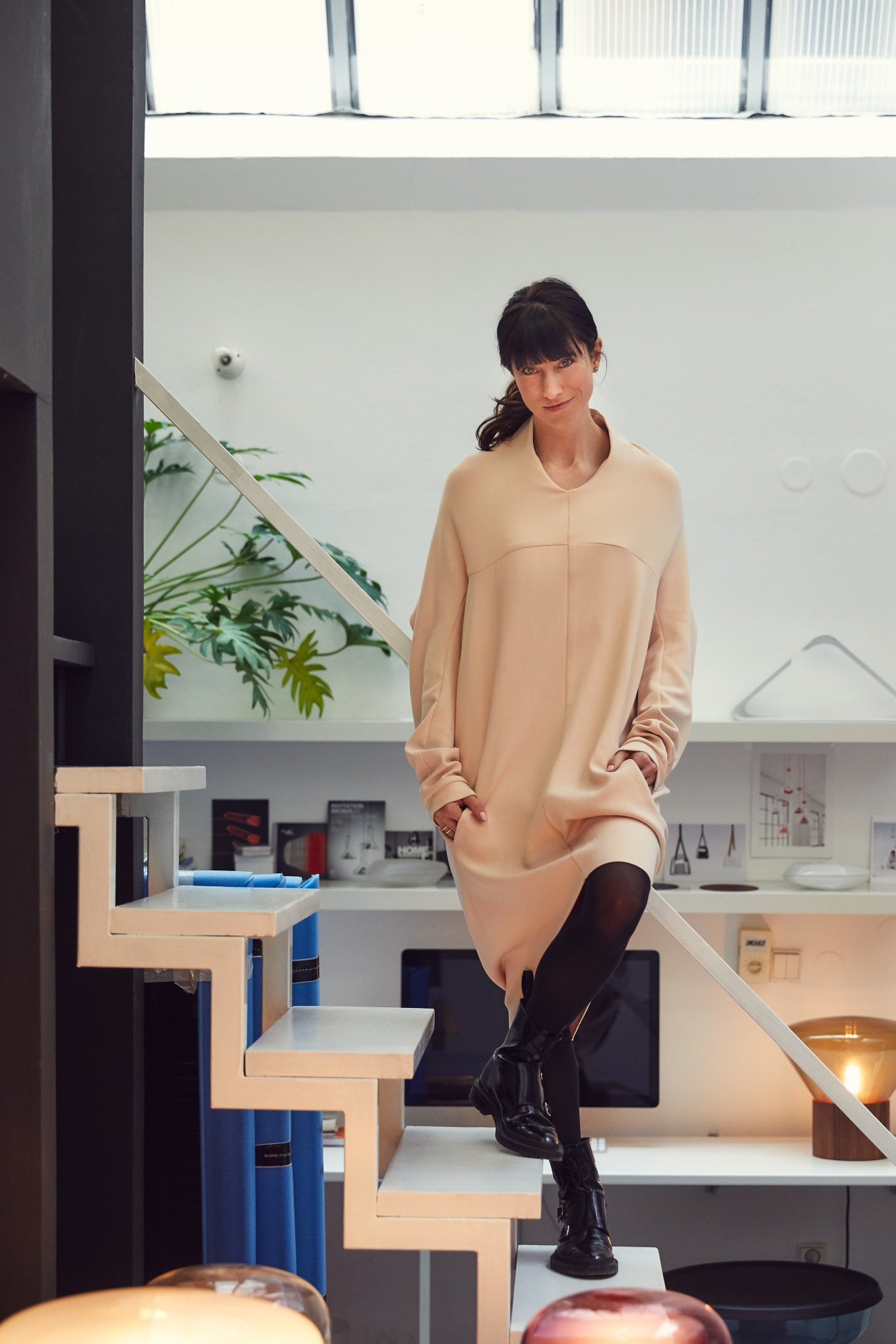 Designer Lucie Koldova wears a beige sweater dress and black boots while posing on modern wooden stairs in a minimalist interior.