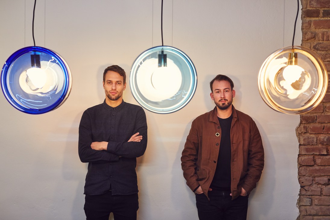 Two men stand against a white wall beneath three colorful pendant lights in blue, light blue, and amber tones.