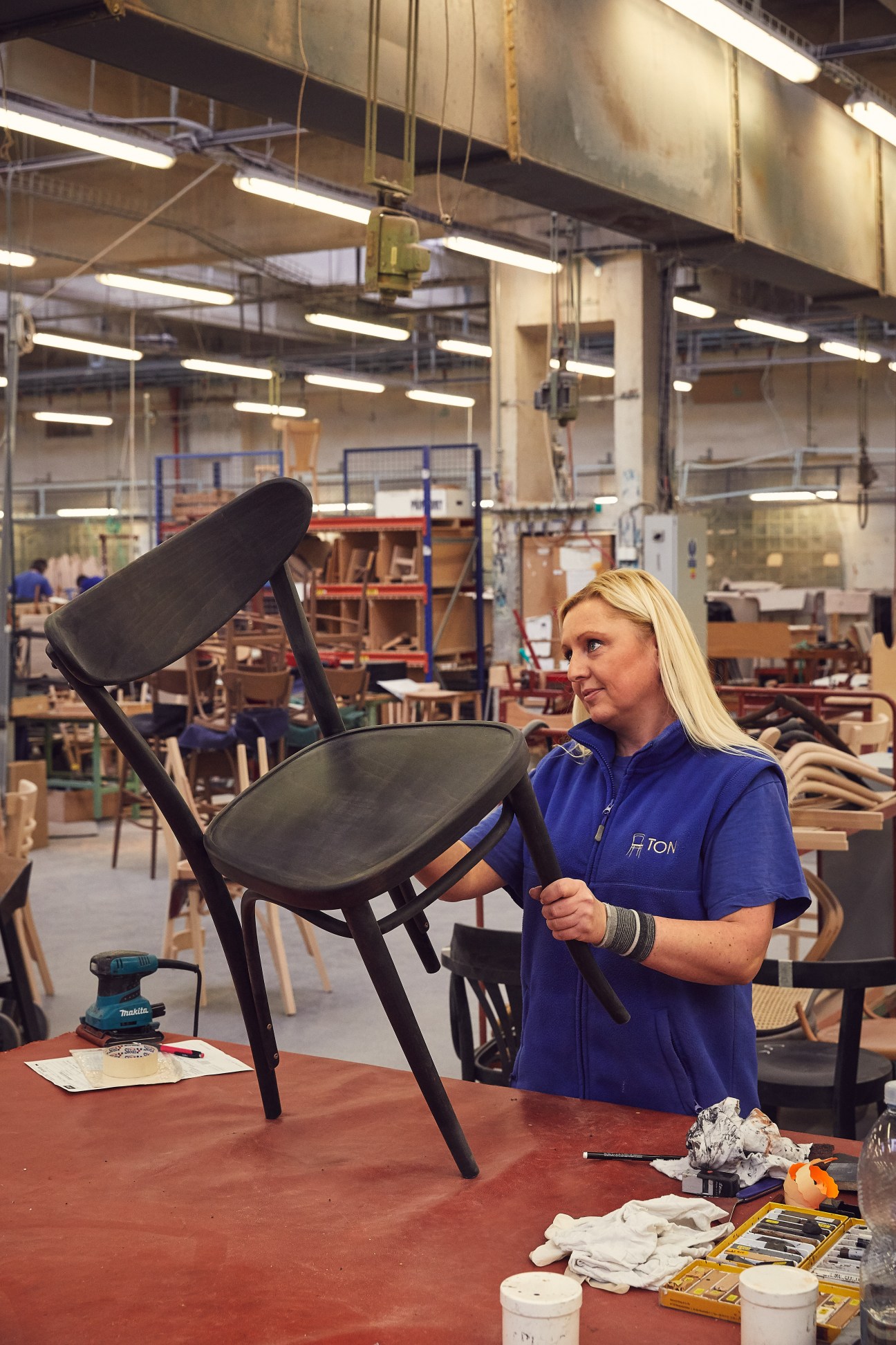 Woman in blue Ton polo shirt inspecting black wooden chair in factory workshop.