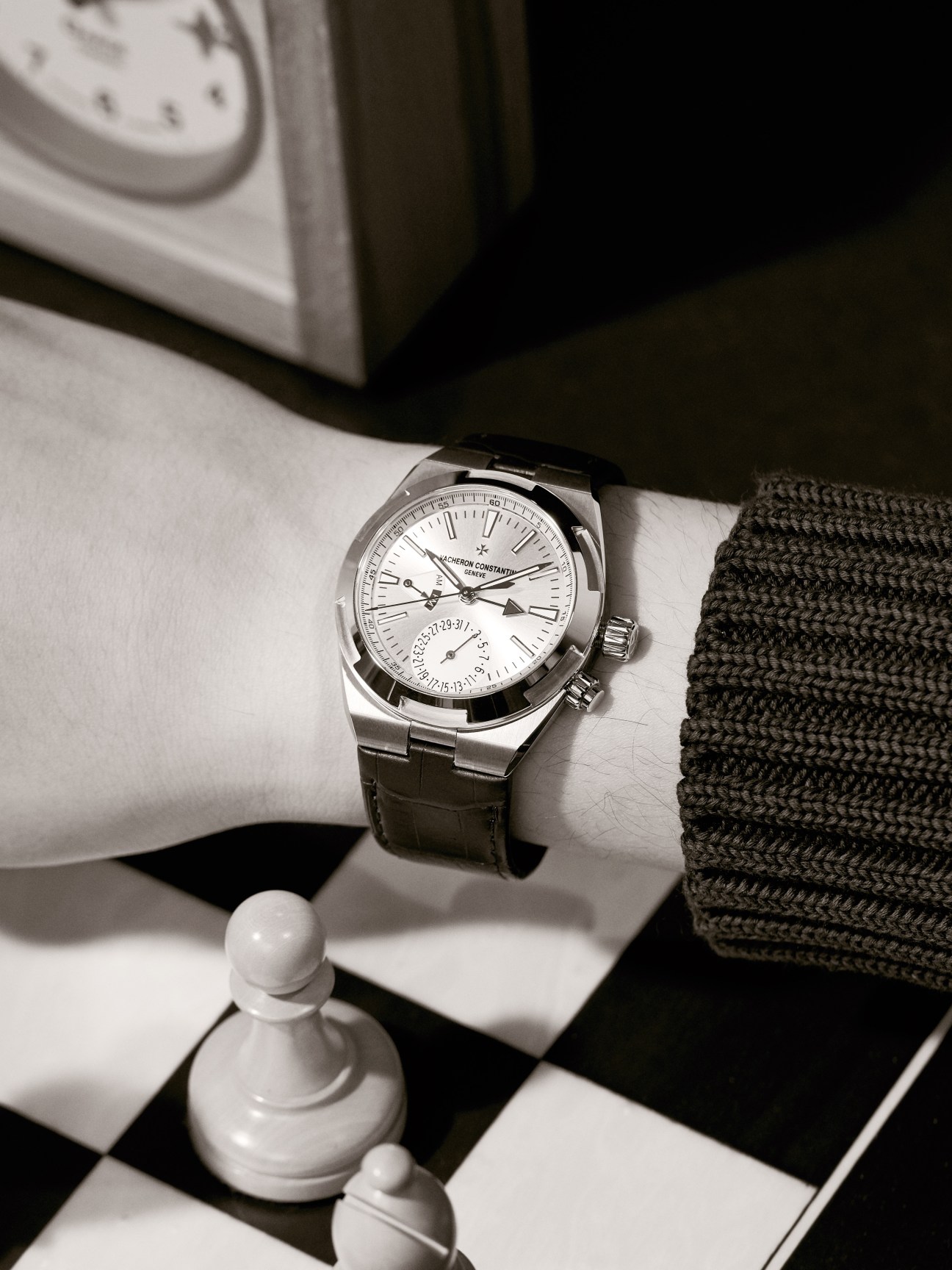 Vacheron Constantin Overseas Dual Time watch worn on wrist with dark jumper against checkered surface.