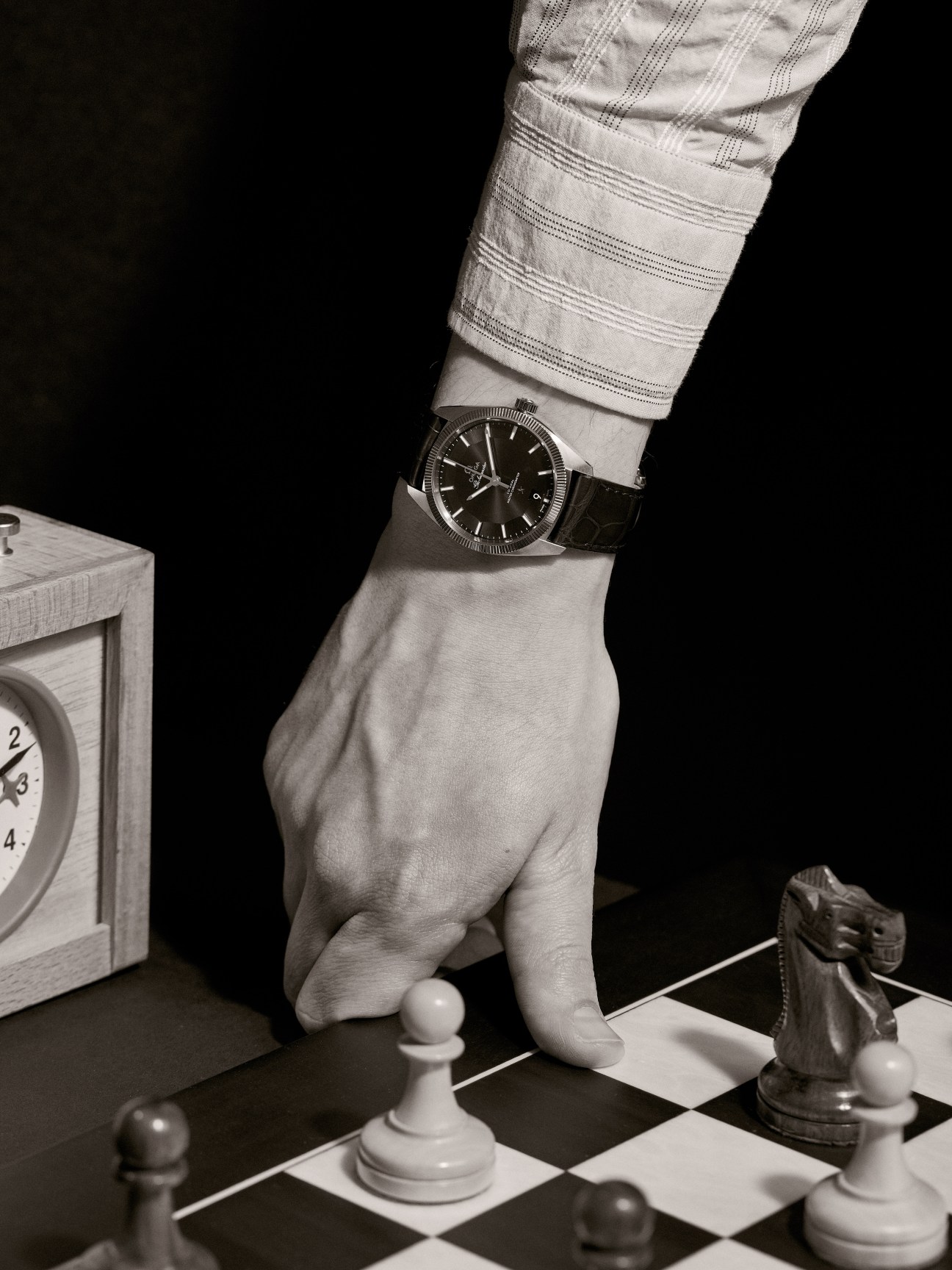 A man's wrist wearing an Omega Globemaster watch over a striped Bagutta shirt, positioned above a chess board.