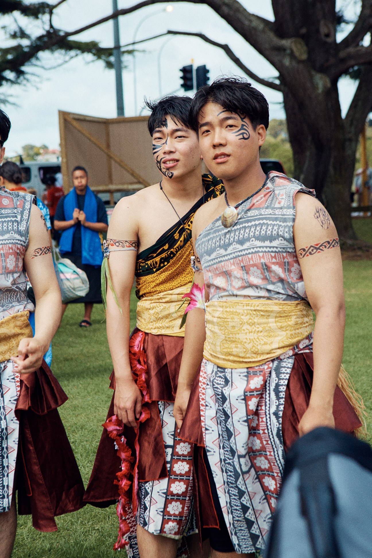 Two young men with traditional Māori facial tattoos and patterned traditional clothing at an outdoor gathering.