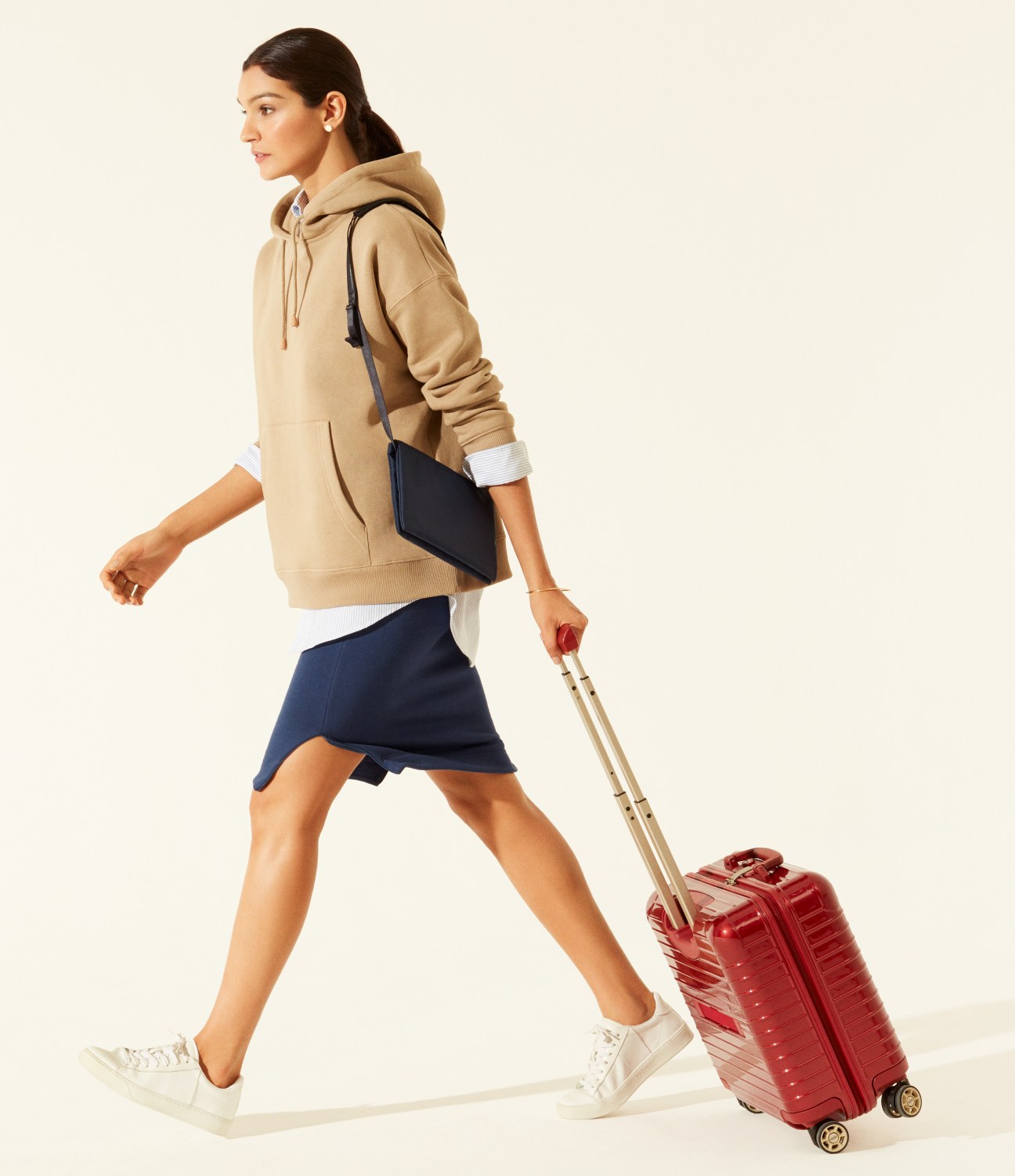 Woman in beige Peak Performance hoodie and navy skirt walking with red Rimowa suitcase.
