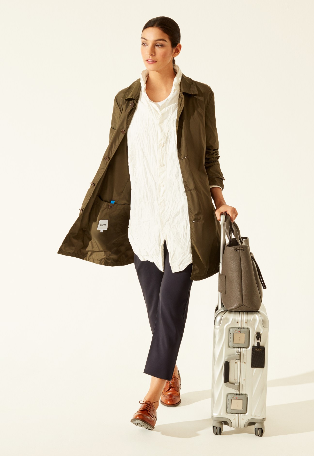Woman in olive Aspesi coat and white Casey Casey shirt carrying Tumi suitcase and bag.