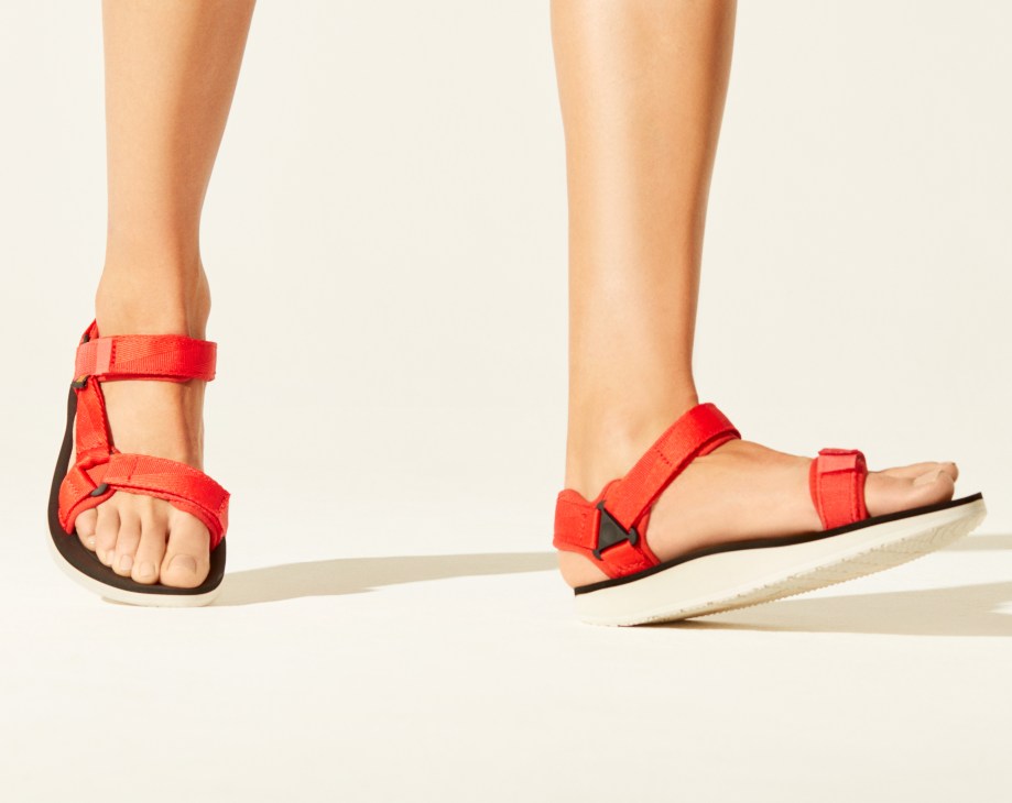 Red Teva sandals with velcro straps and dark soles on bare feet against a neutral background.