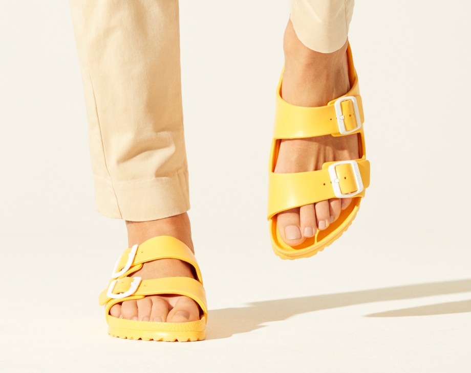 Model wearing cream trousers and yellow Birkenstock sandals with white buckles against a white background.
