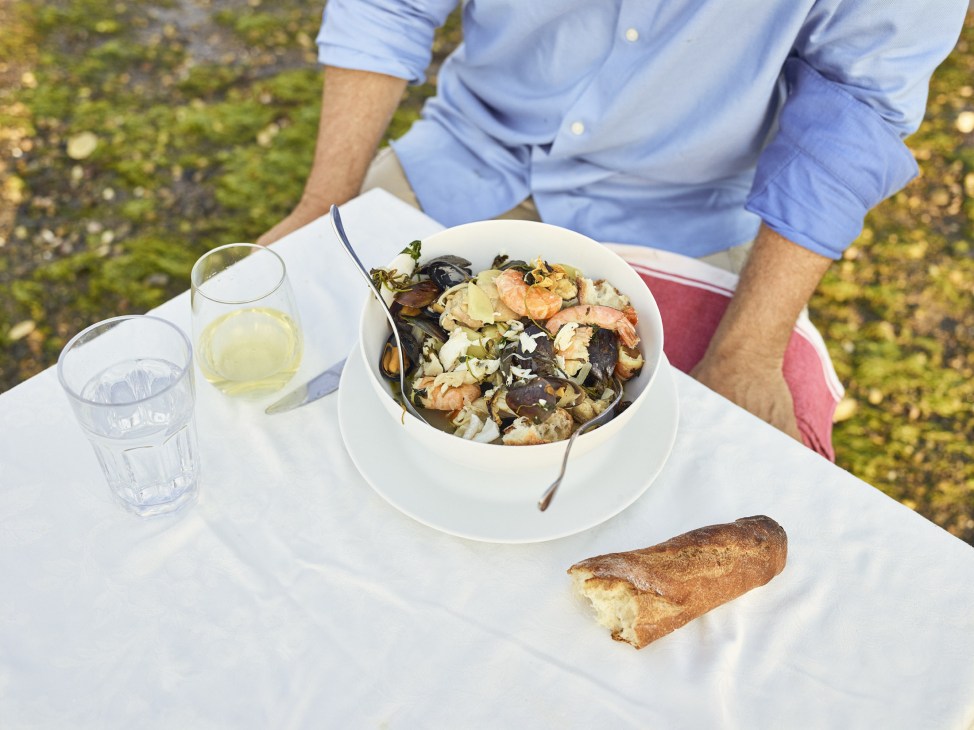 Man in blue shirt serving Chilean fisherwoman's stew of seafood and chicken in white bowl at outdoor table.