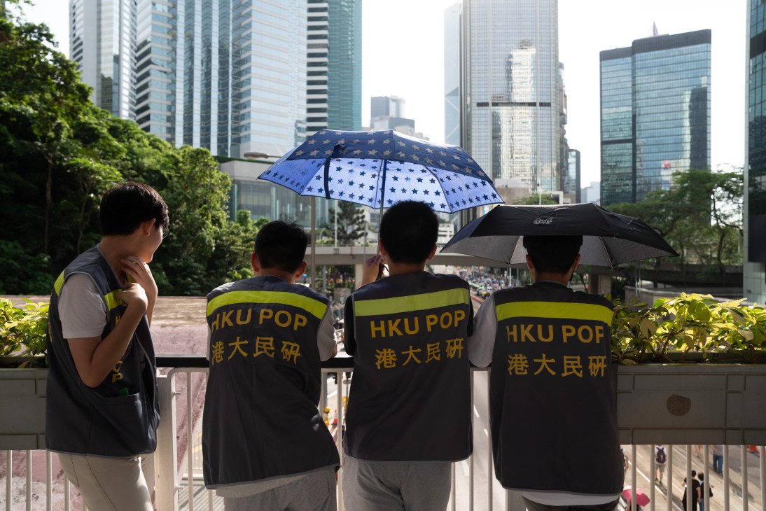 Hong Kong University PopStudies team members wearing yellow and black vests hold umbrellas during a protest march with city skyscrapers in background.