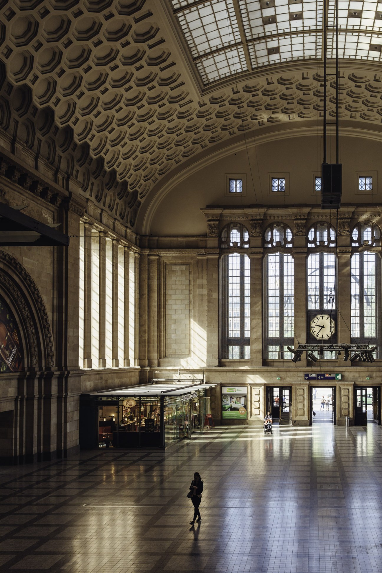 Interior of Leipzig's main train station with ornate coffered ceiling, clock, and a single figure walking across the floor.