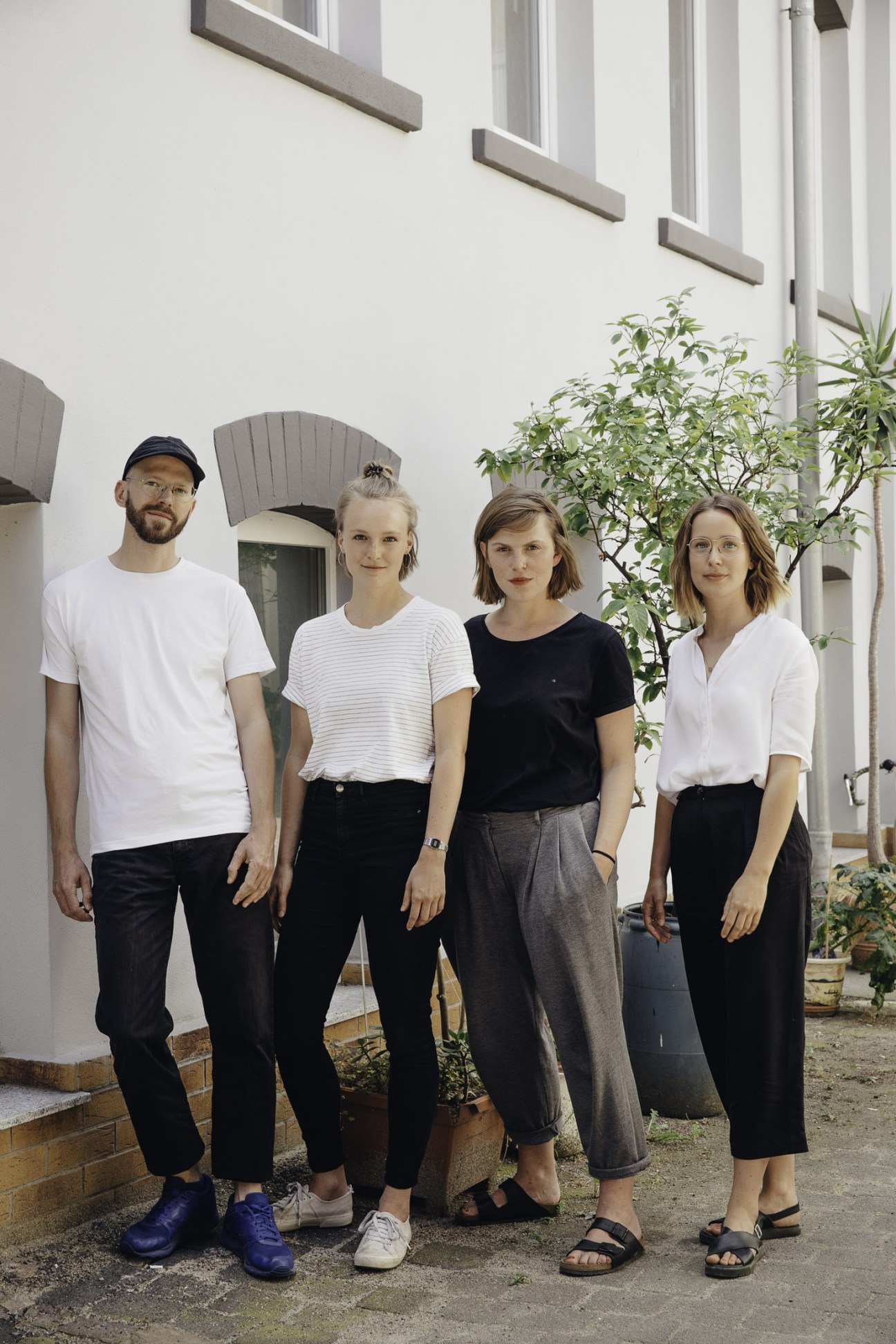 Four graphic designers stand outside a white building with large windows and green plants.