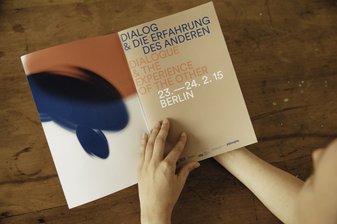 Hand holding a design publication with bold orange and blue geometric shapes on wooden surface.