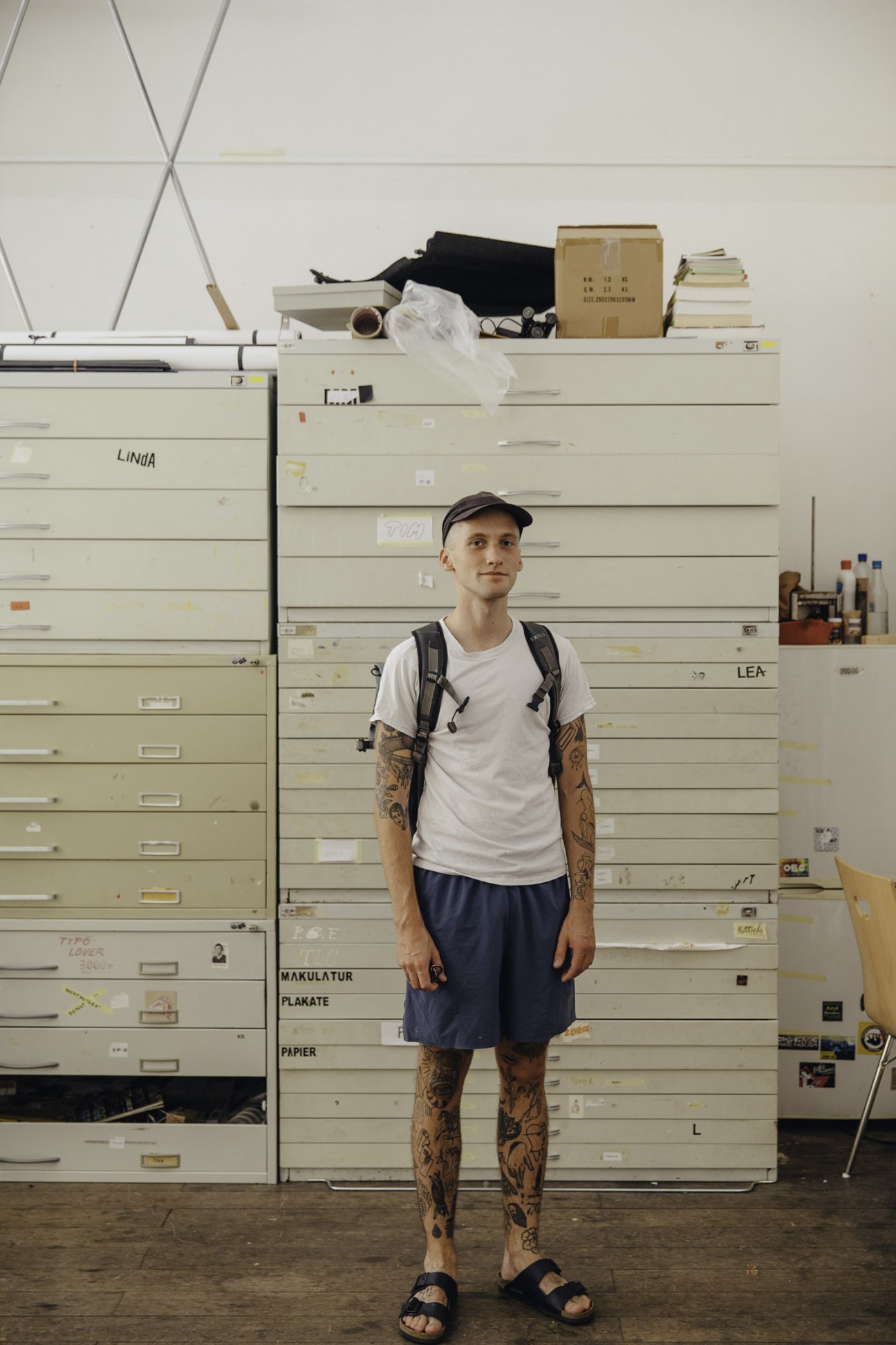 A young man with tattooed arms stands in front of industrial shelving units filled with papers and materials in a studio space.