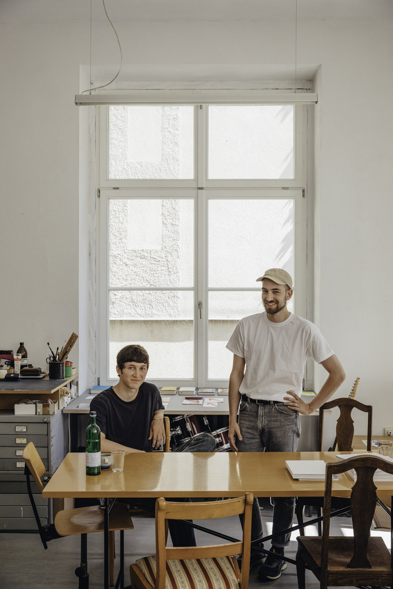 Two men stand at a wooden table in a bright studio with large windows; one seated wearing a black shirt, one standing in a beige cap and white t-shirt.