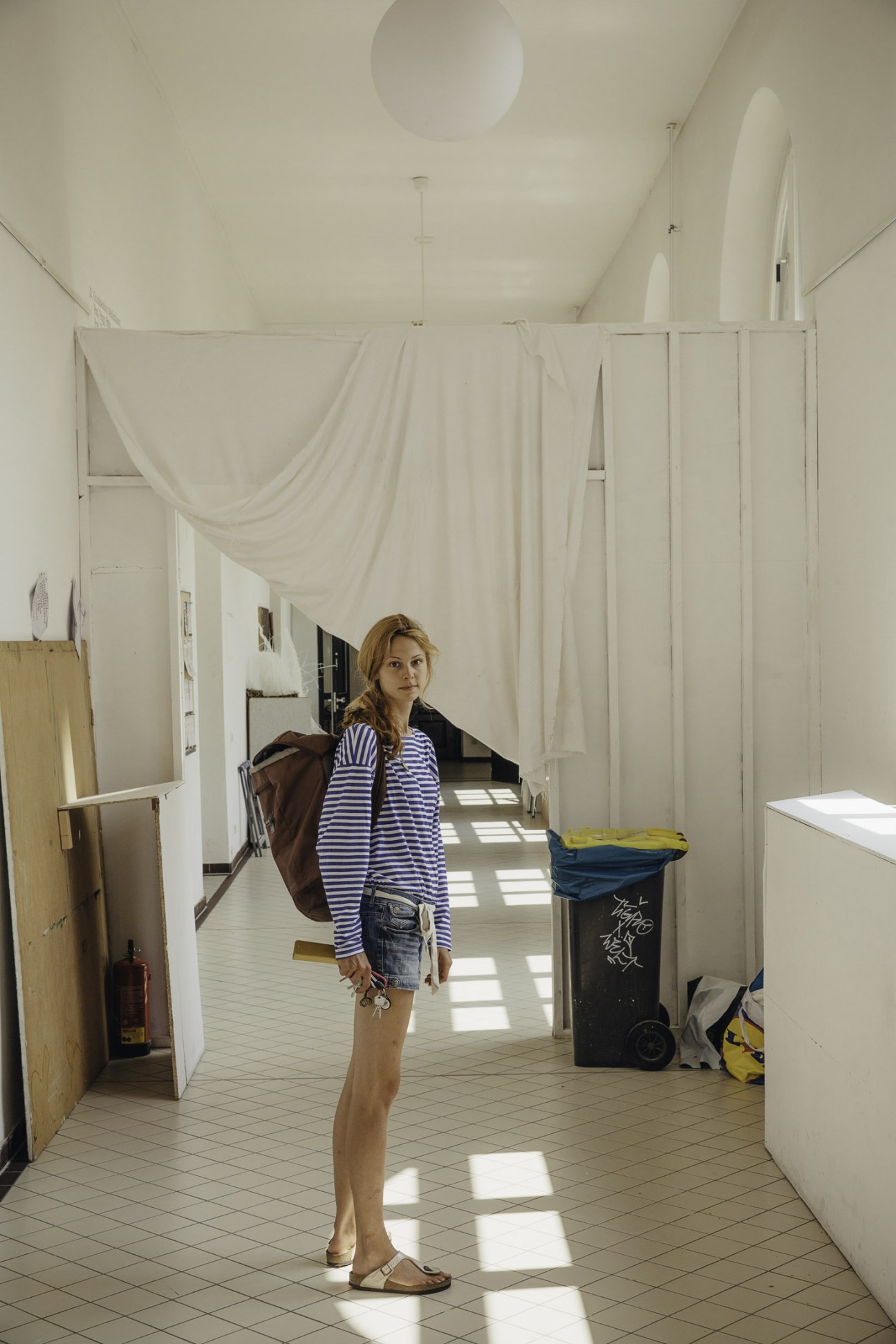A woman in a striped shirt stands in a bright hallway with white curtains and industrial fixtures.