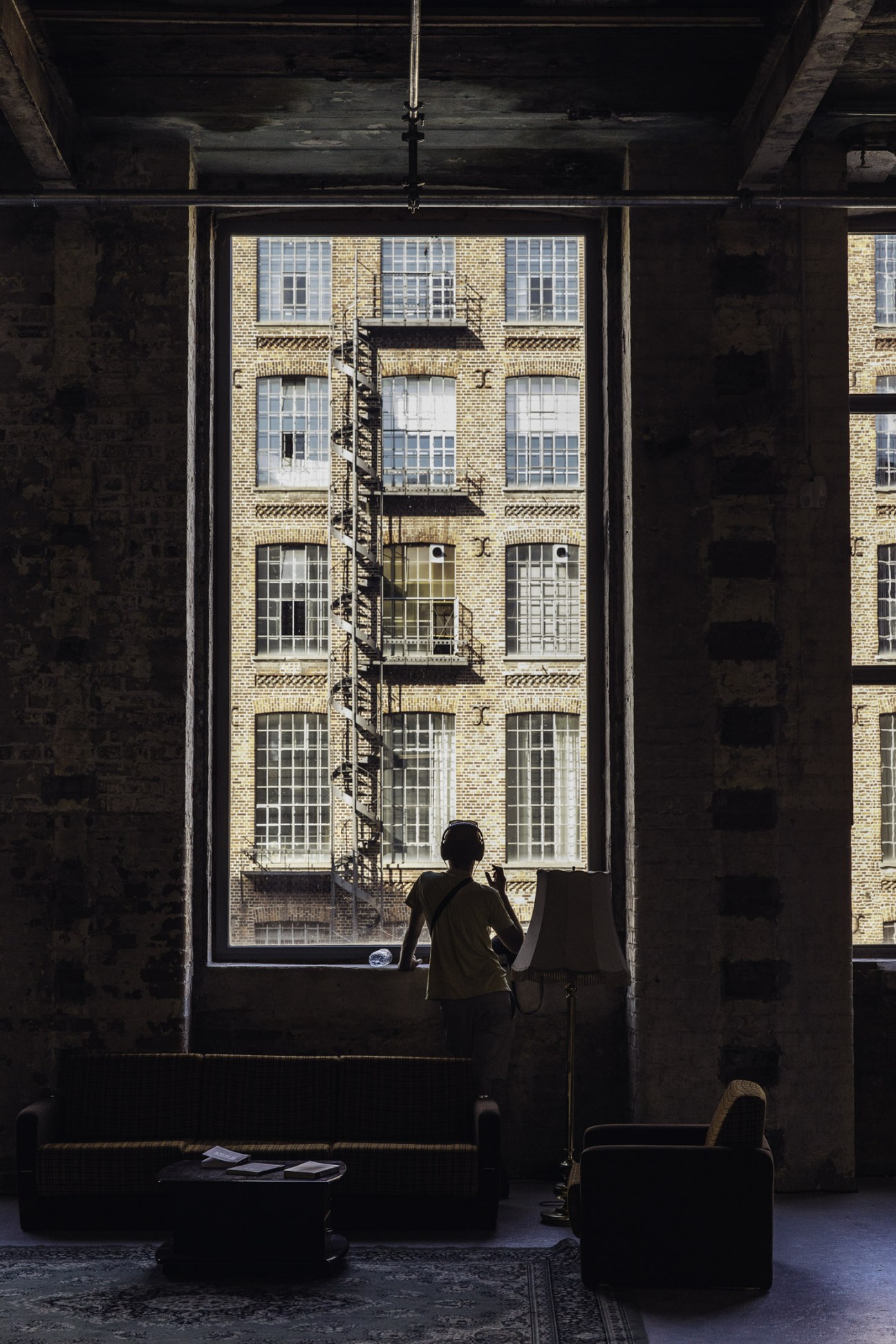 Person standing at large window looking out at brick building facade with metal fire escape stairs.