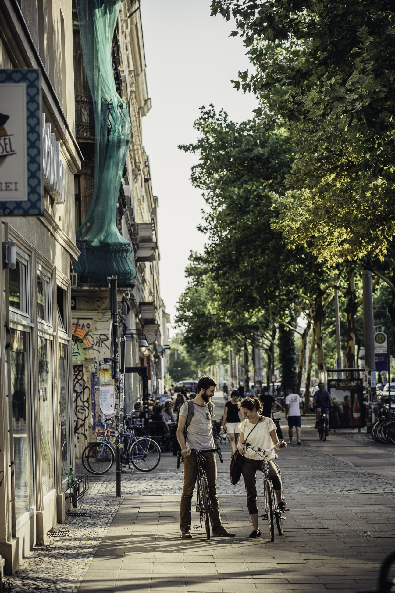Two cyclists ride side-by-side on a tree-lined Leipzig street with historic buildings and storefronts.