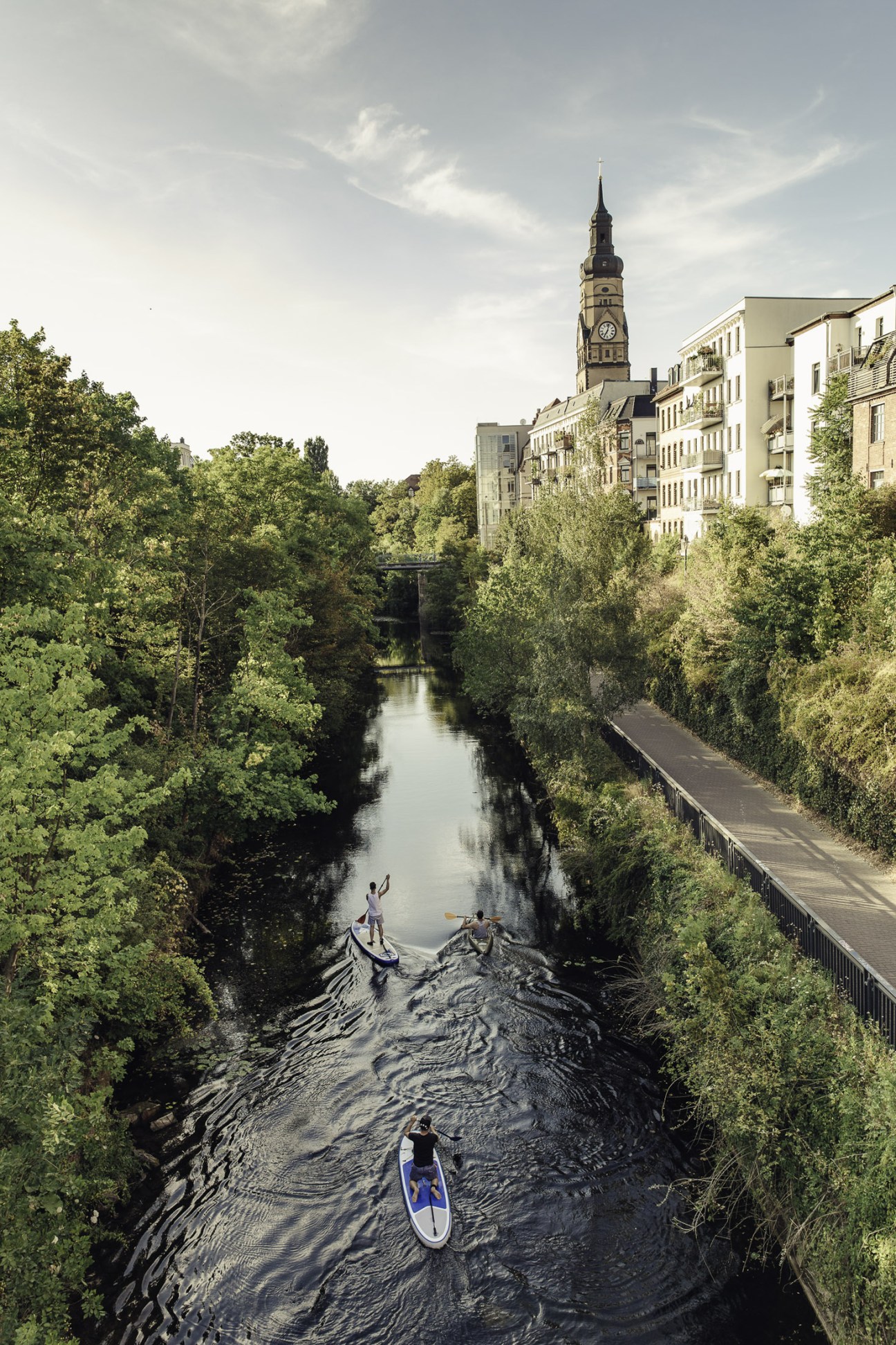 People paddleboarding and wading in a river flowing through Leipzig with historic buildings and a church spire visible in the distance.