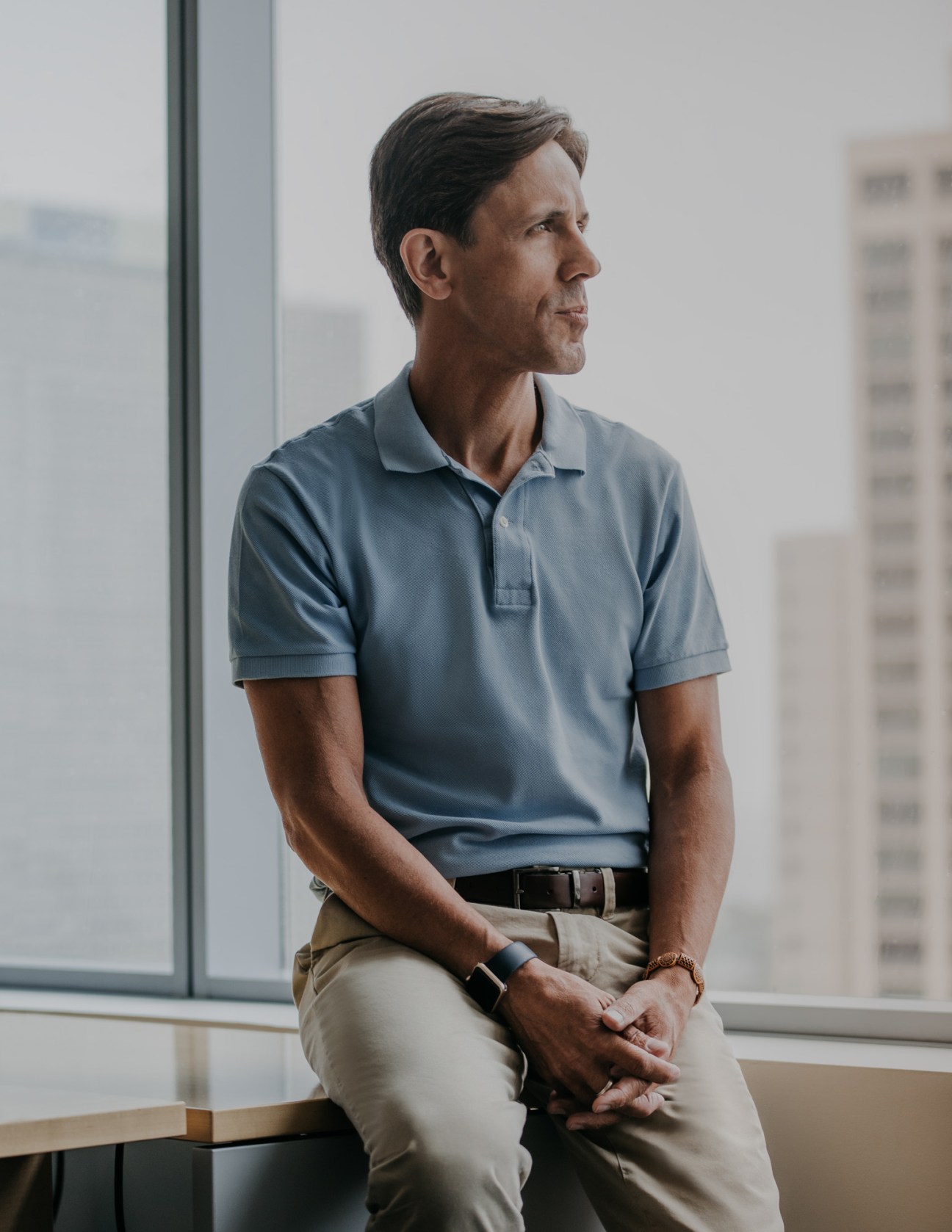 Man in blue polo shirt and khaki trousers sitting by a window overlooking Chicago skyscrapers.