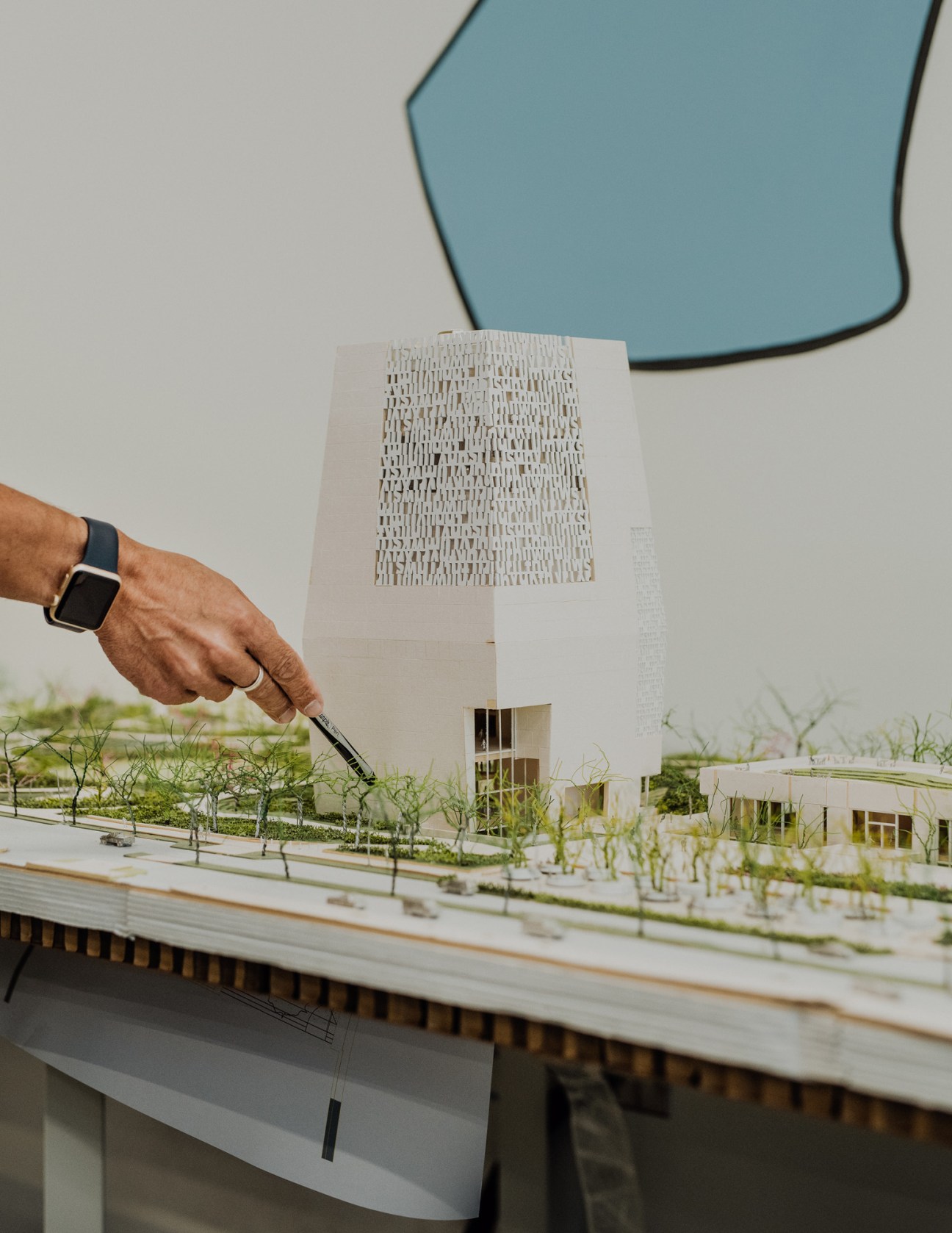 A hand points to a white architectural scale model of the Obama Presidential Center campus with a blue-tinted mirror visible above.