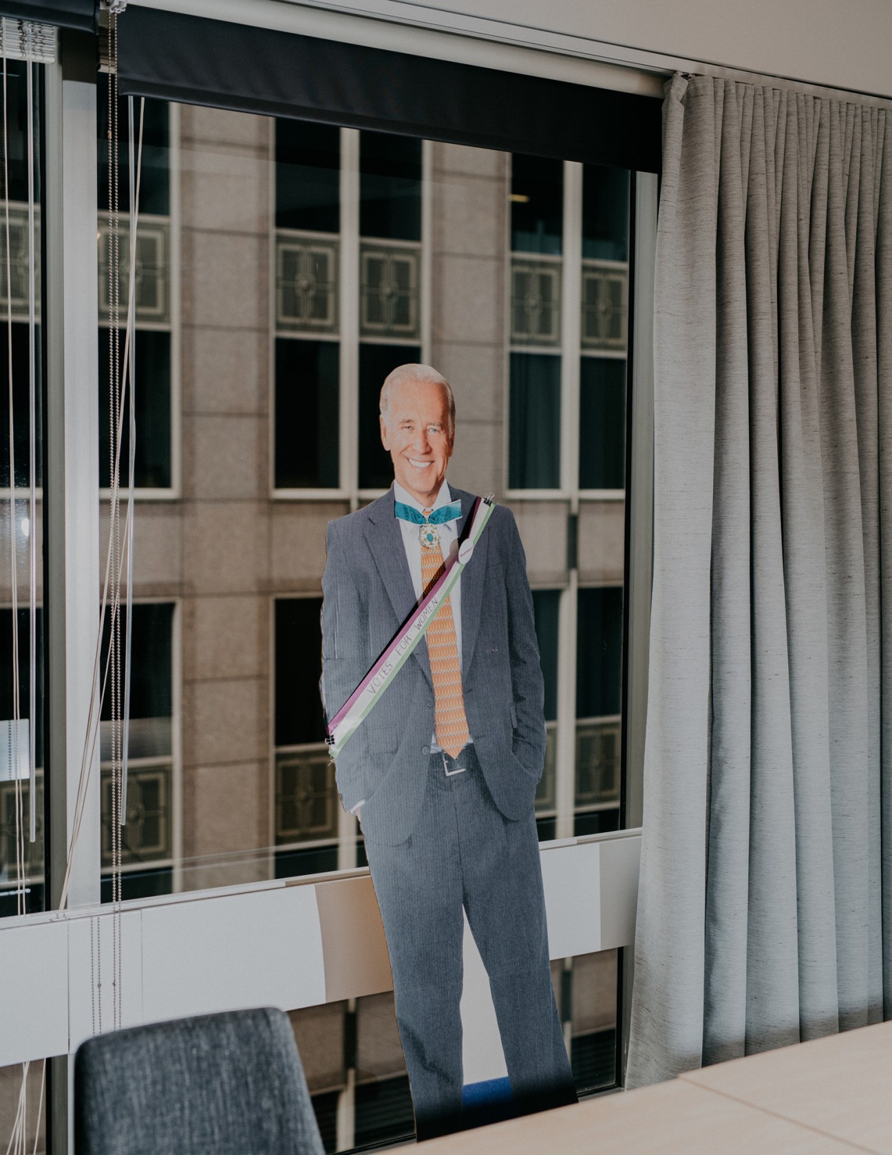 Cardboard cutout of Barack Obama wearing a gray suit and colorful sash displayed in an office window.