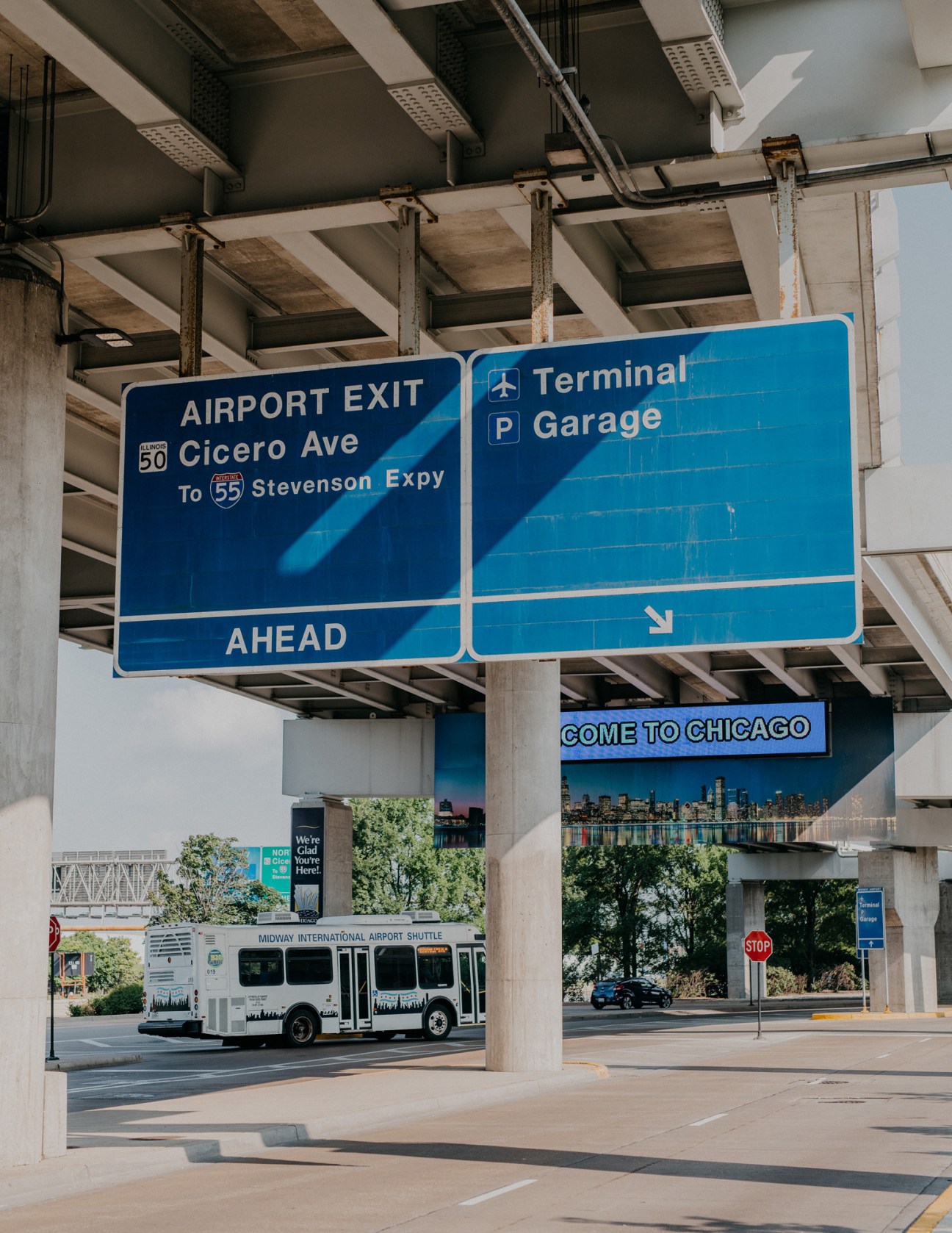 Blue directional signs under a concrete overpass at Midway Airport in Chicago, with a shuttle bus and skyline visible below.