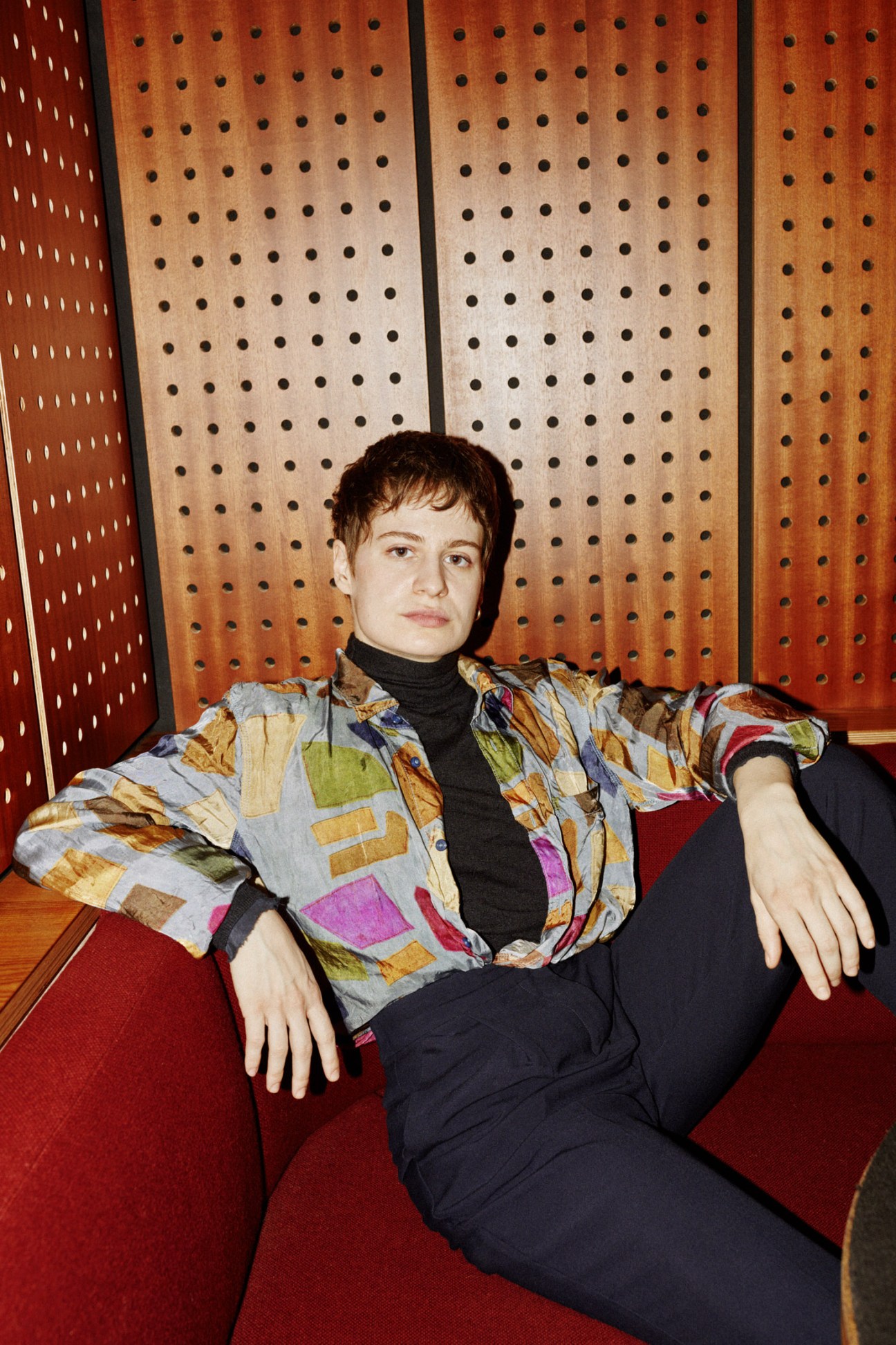 Christine and the Queens seated on a red chair wearing a patterned jacket and black turtleneck