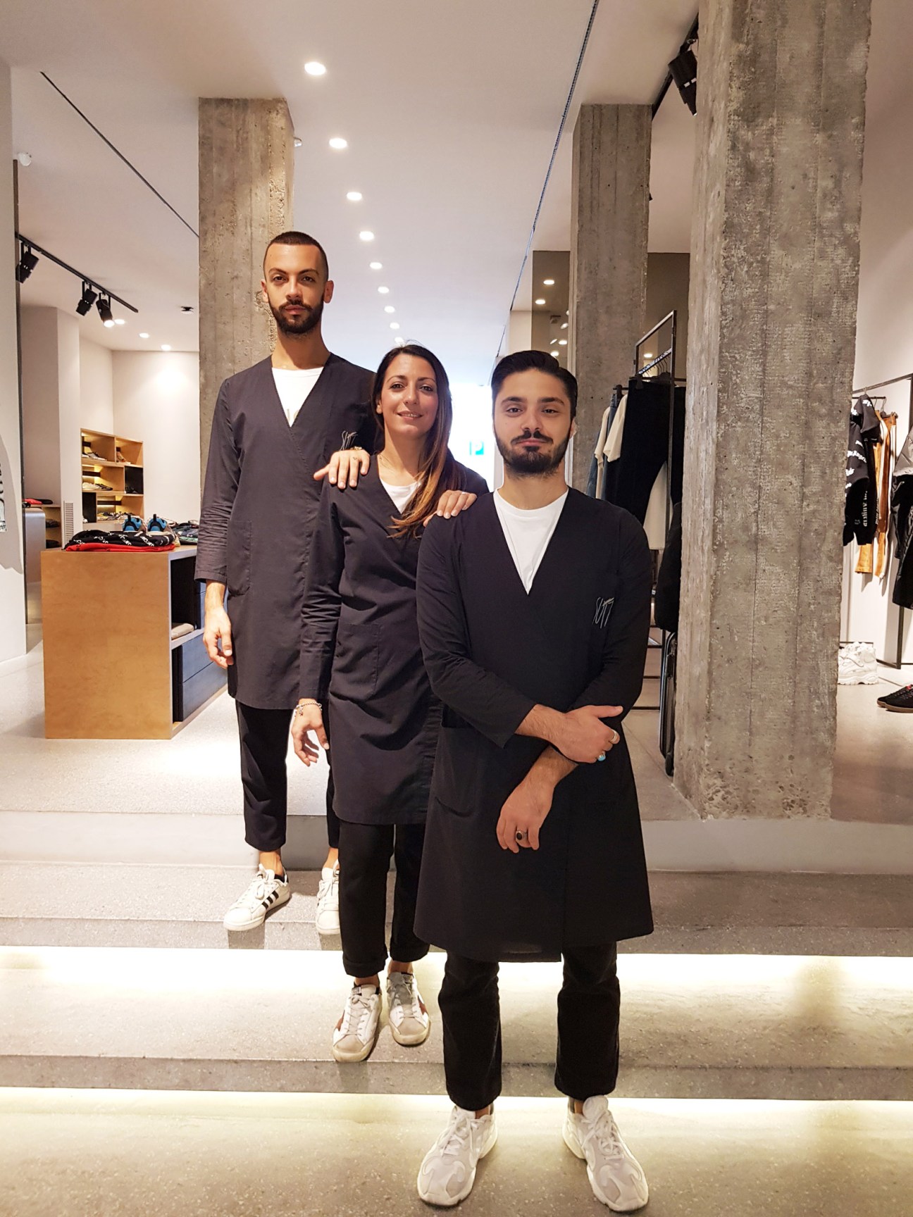Three staff members pose together inside Suit boutique in Lecce, a luxury fashion shop with concrete pillars and modern retail displays.