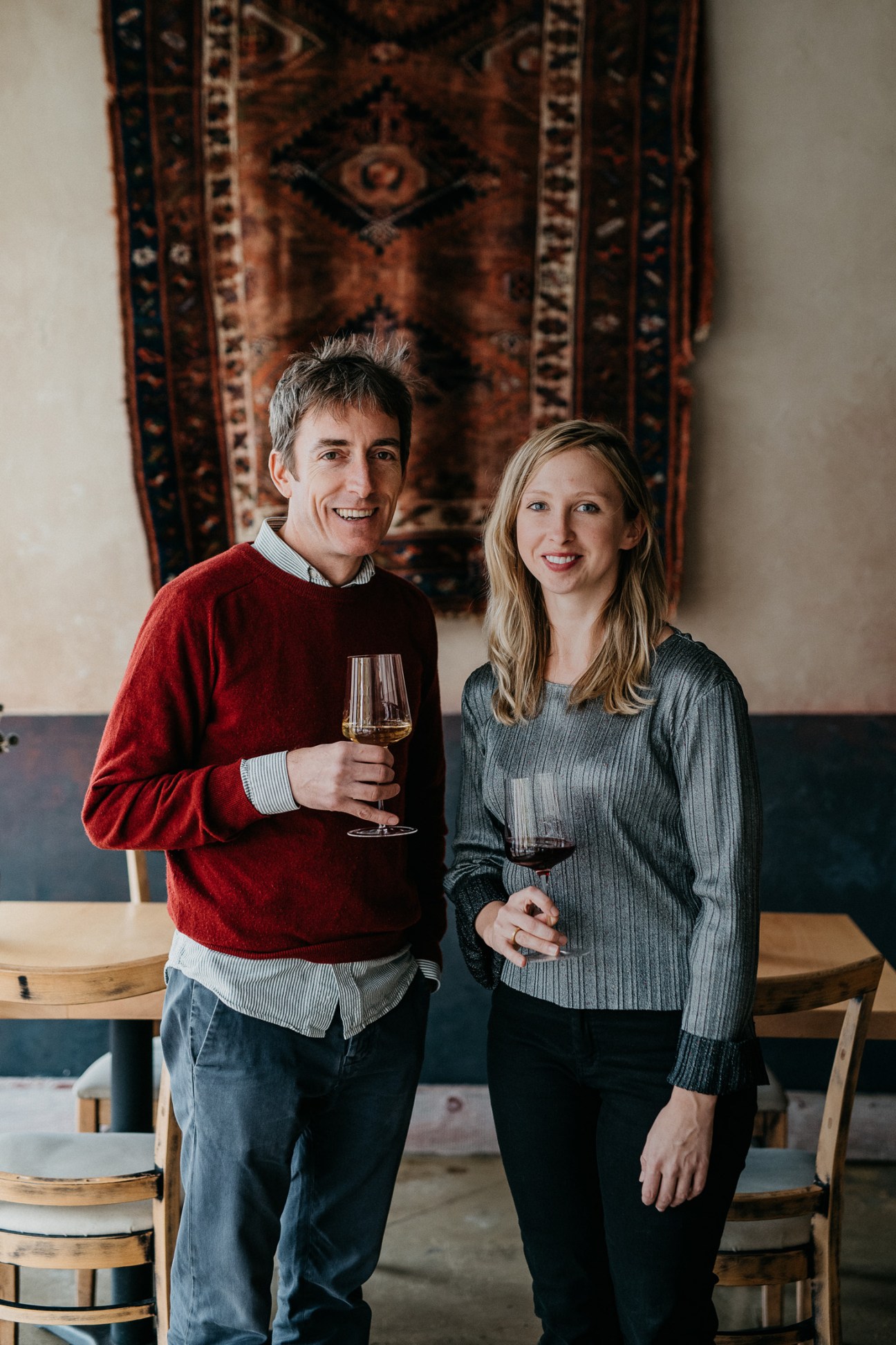 Two restaurant owners pose with wine glasses in front of a decorative textile backdrop.