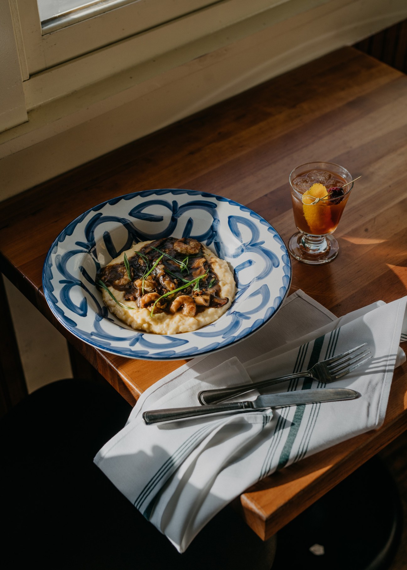 Grits dish with mushrooms and greens served in decorative blue and white bowl alongside cocktail