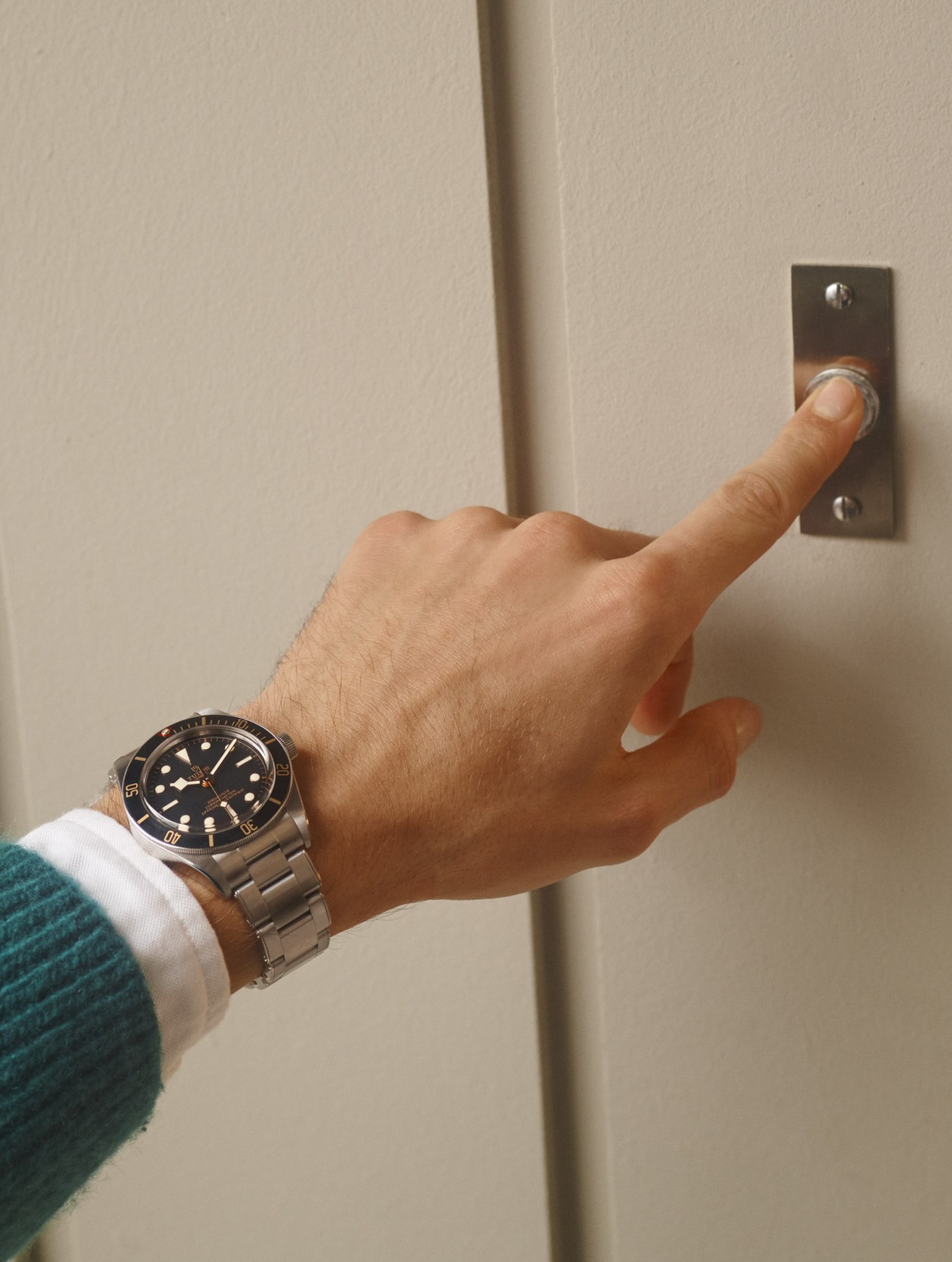 Man's hand wearing Tudor watch and white shirt cuff pressing a door buzzer.