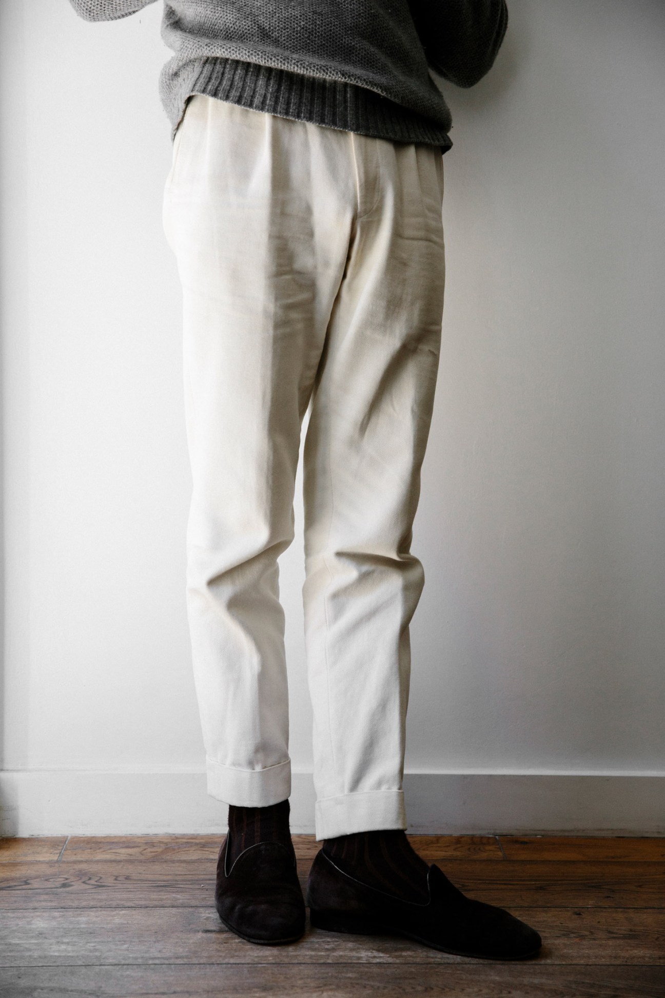 Model wearing cream pleated trousers with rolled cuffs and black suede ankle boots.