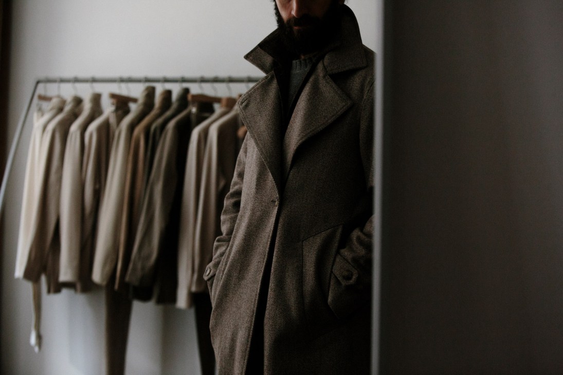 Man wearing Stòffa's asymmetric coat standing in front of rack of neutral-toned garments.